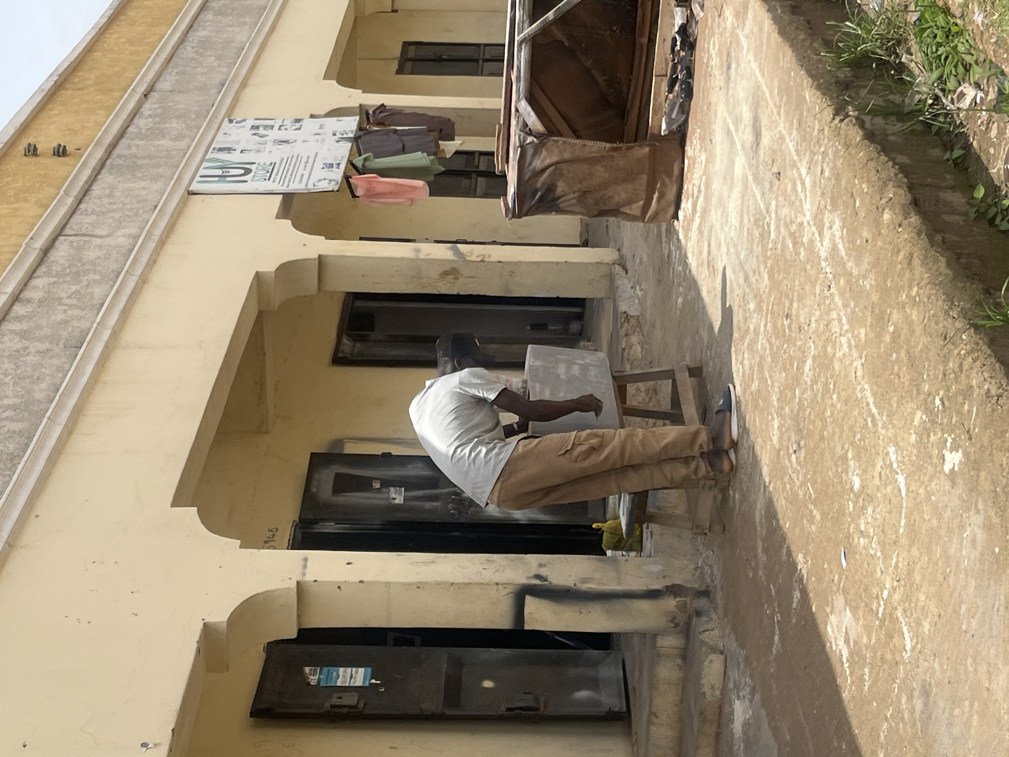 A man is working on a large, light-colored object, possibly stone, on a wooden stool in front of a building in Bukuru, Nigeria. The building is painted yellow with arched doorways and windows. A sign for "HUY STORE" hangs above the entrance. The ground is a mix of concrete and dirt, with some grass and debris visible. The man is wearing a light-colored shirt and tan cargo pants. He is focused on his work, with his head bent down. The scene appears to be an outdoor workshop or market area.