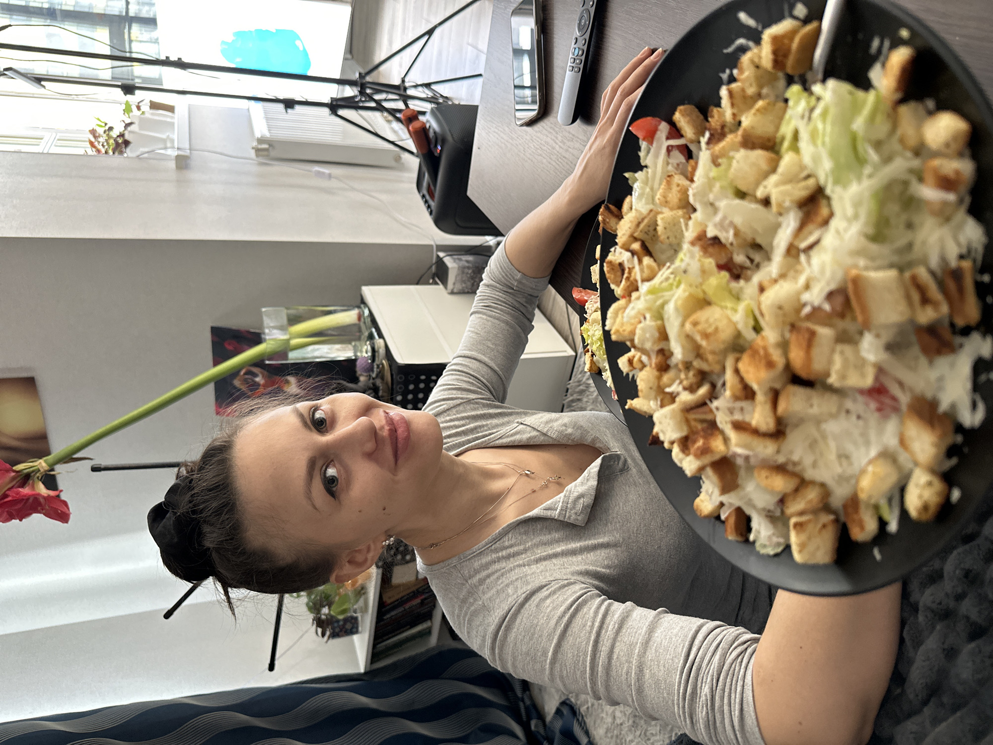 A woman sits at a table inside a room, holding a large plate of salad. She wears a grey long-sleeved shirt and a necklace. Her hair is pulled back. The salad is on a black plate and appears to contain lettuce, croutons, and tomatoes. On the table near her hand are a remote control and a smartphone. Behind the woman is a shelf with books and other items. A flower with a long green stem is to the left of her. In the background, there is a window with a lighting setup. The city of Unknown, Unknown is not discernible from this image.