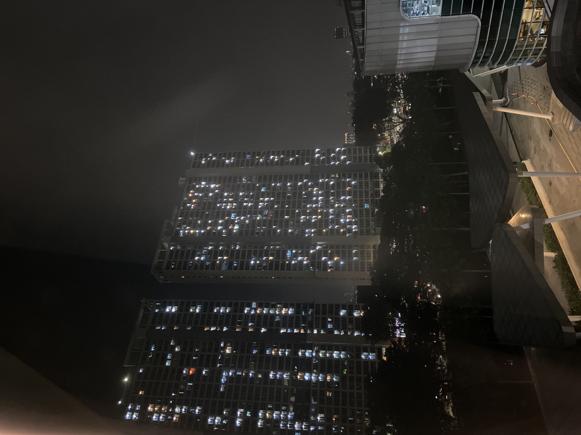The image depicts a night scene in North Jakarta, Indonesia. Tall buildings with illuminated windows dominate the view, suggesting a densely populated urban area. The sky is dark and possibly overcast. Trees are visible amidst the buildings, and a modern structure with curved architectural elements is situated on the right side of the frame. The overall atmosphere is dimly lit and suggestive of a quiet, late-night setting in the city.
