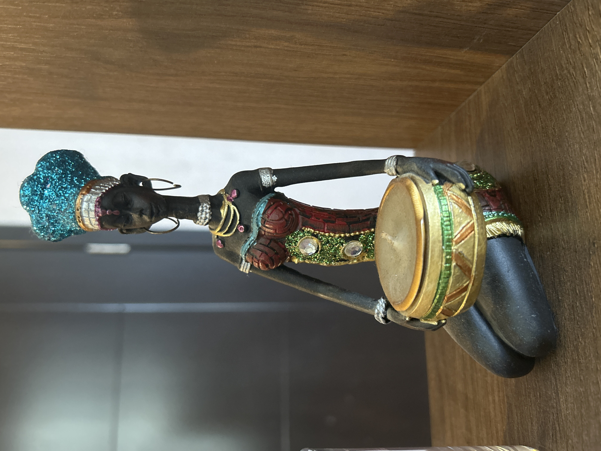 The image displays a close-up, eye-level shot of a decorative figurine of a kneeling woman, situated on a wooden surface in a location identified as Unknown, Unknown. The figurine, which appears to depict a woman of dark complexion, is adorned with a bright, glittery turquoise headwrap featuring a gold and pink banded trim. Large gold hoop earrings are visible, along with multiple gold necklaces, a silver choker, and a prominent pink gemstone on the chest. The woman wears a reddish-brown bodice with textured patterns, accented with green glittered sections containing three large, clear circular rhinestones on her left side (viewer's right). Silver bands encircle her upper arms and wrists. She holds a circular, drum-like object, predominantly gold with green and beige geometric patterns, suggestive of a candle holder given the visible light-colored wick on its top surface. The wooden surface beneath the figurine exhibits a subtle grain pattern. The background on the left consists of a smooth, dark grey wall or panel, while the right side features a vertical wooden panel matching the horizontal surface.