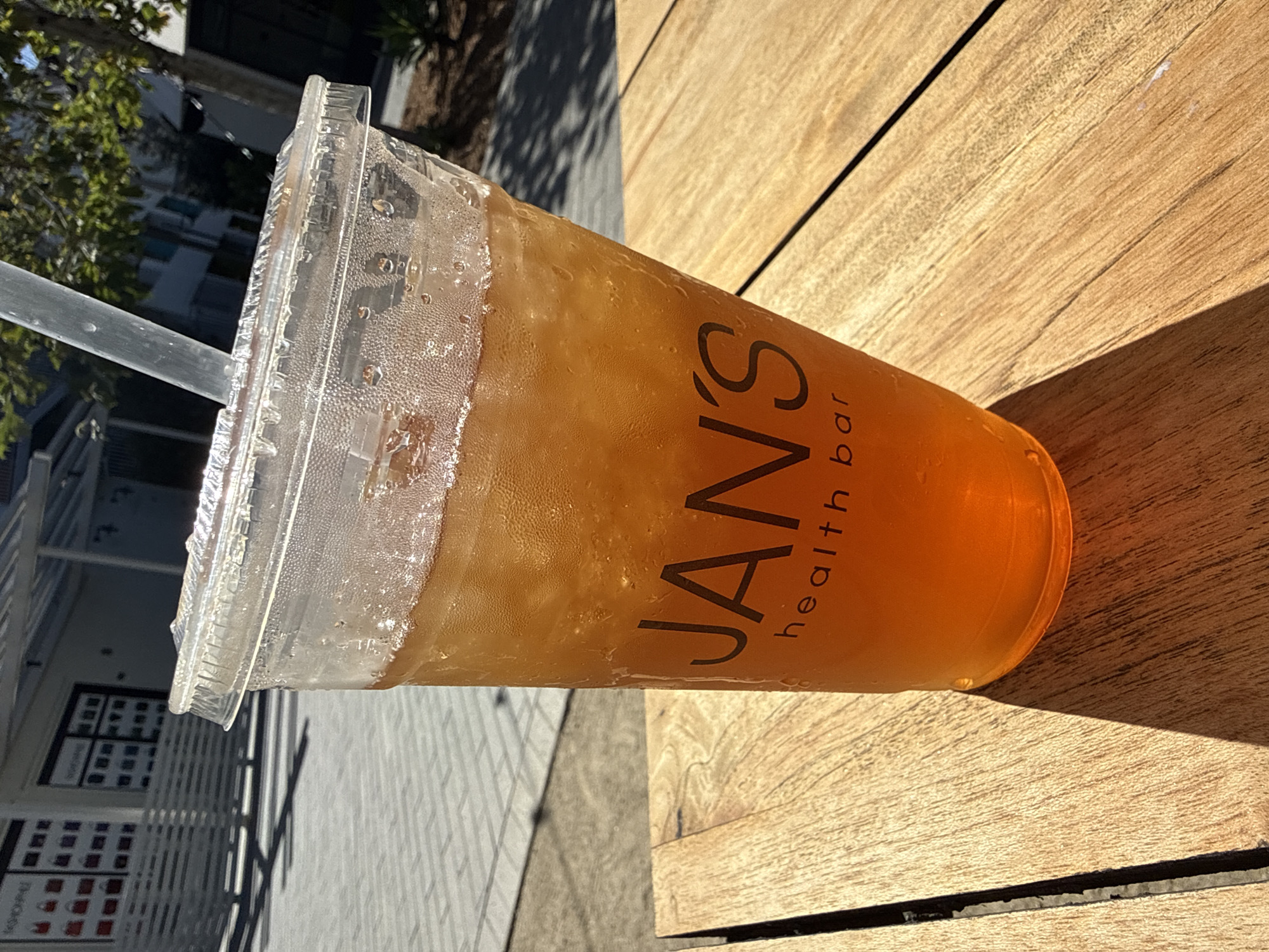 An eye-level shot captures a clear plastic cup filled with an amber-colored, iced beverage, likely iced tea, sitting on a light-colored slatted wooden table. A plastic straw is inserted into the drink through the lid. Condensation beads are visible on the exterior surface of the cup and lid, indicating the drink is cold and the day is warm.
Prominently displayed on the side of the cup is the text "JAN'S health bar" in a dark, stylized font. Strong shadows cast across the wooden table suggest bright, direct sunlight, indicating it is daytime. In the background, out of focus, are glimpses of urban architecture, including tiled pavement, buildings with windows, and some green foliage from trees. The scene suggests an outdoor setting, possibly a cafe patio or seating area in San Diego, United States of America, during a clear, sunny day. The overall impression is one of a refreshing drink enjoyed outdoors.