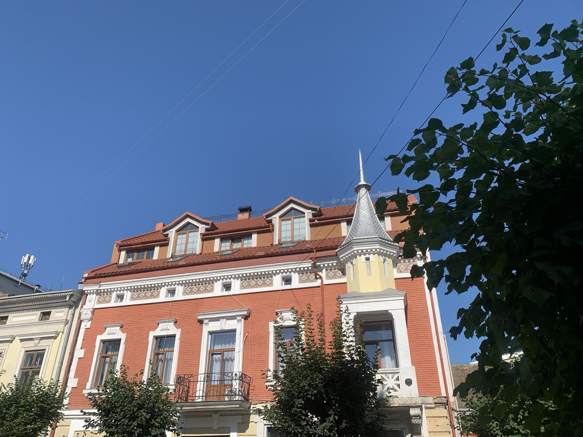 The image shows a close-up of the upper facade of a red brick building with a distinctive yellow turret topped by a silver conical roof. The building has multiple windows with brown frames and white trim, some of which are adorned with decorative architectural elements. A small balcony with black wrought-iron railings is visible. Above the red brick, there is a white decorative band with a repeating circular pattern. The building has a red tiled roof with several dormer windows. To the left, a section of a yellow building with similar architectural details is partially visible. In the foreground, dark green foliage from a tree partially obscures the view of the buildings. The sky above is a clear, bright blue with two thin white lines stretching across it, likely power or telephone lines. A cell tower is visible on a neighboring building to the far left. The building depicted is likely located on Svobody Street in Kolomyia, Ukraine, given its architectural style. There are no people visible in the image.
