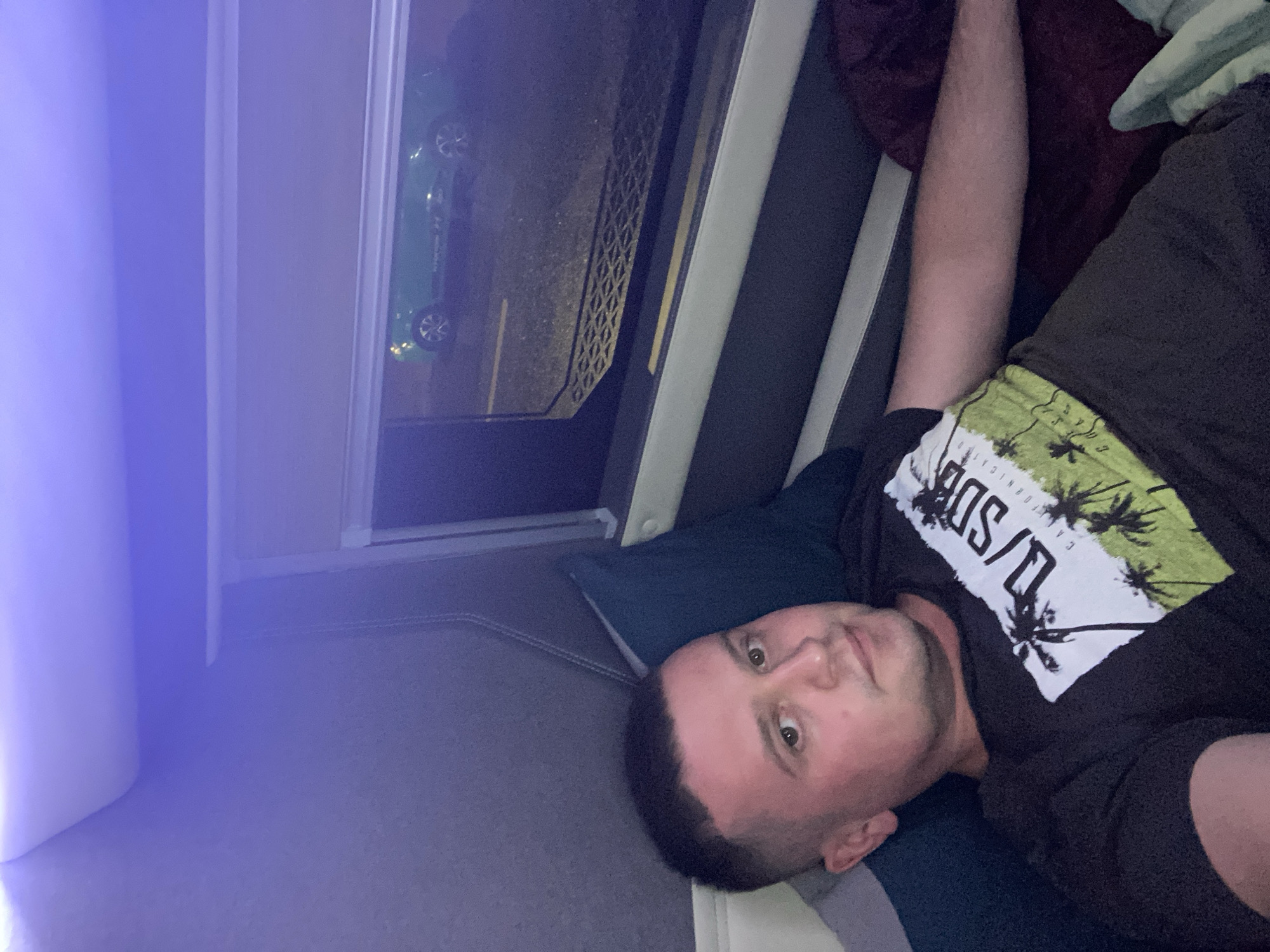 The image shows a man lying down, looking towards the camera. He is wearing a black t-shirt with a graphic design featuring palm trees and text. His head is resting on a dark blue pillow. To his right, a maroon blanket is visible. In the background, through a window, a green car is parked on what appears to be a street or parking lot. The scene is illuminated by a purple light source, likely from an LED strip, located above and to the left of the frame, casting a soft glow on the wall and ceiling. The overall environment suggests an interior setting, possibly a vehicle's sleeping cabin or a small room. There is no information in the image to suggest the location is in Ea Drăng, Vietnam.
