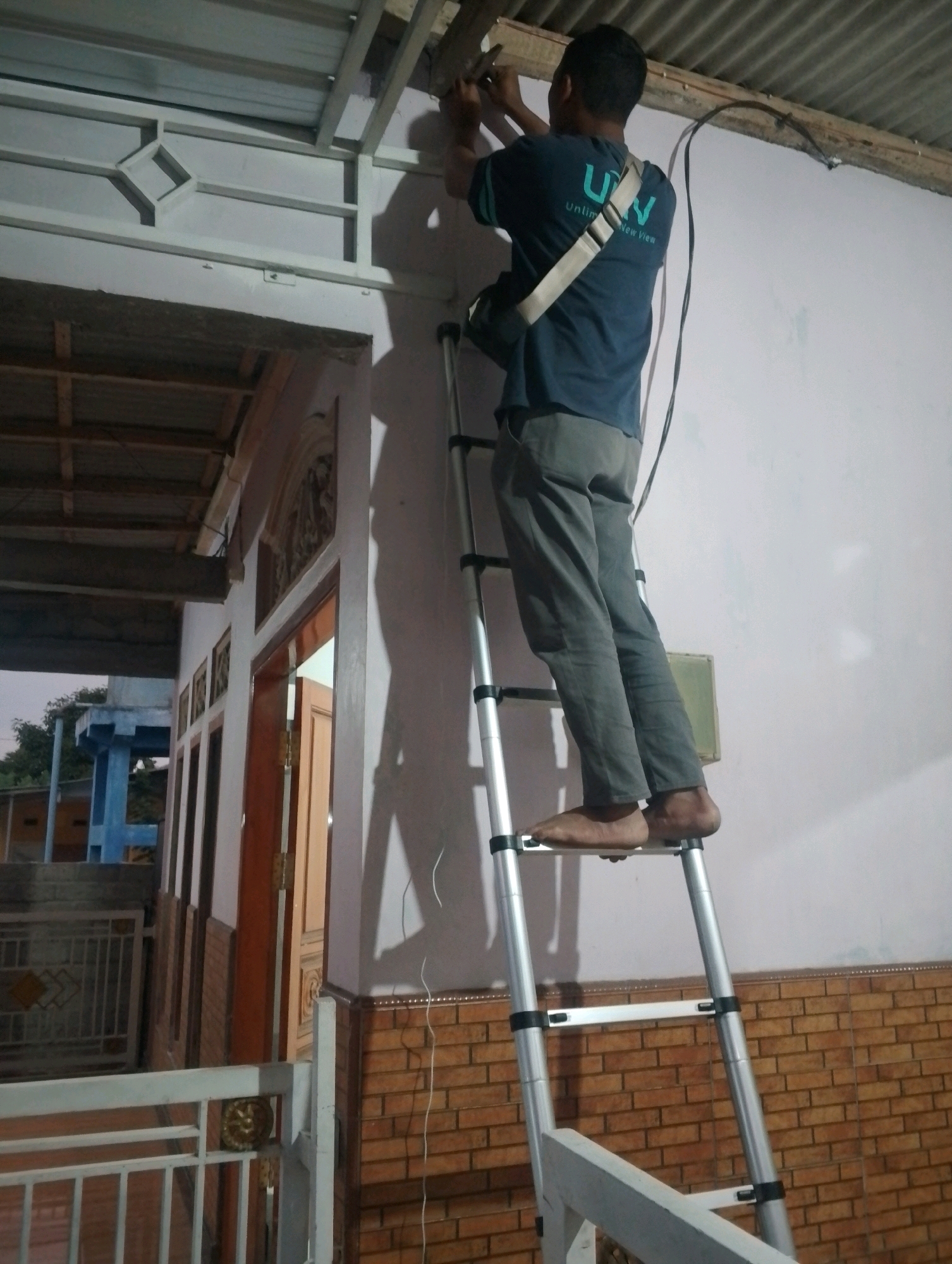 A man is standing on an aluminum extension ladder, working on the exterior of a light pink building, possibly installing or repairing an air conditioning unit. The man is wearing a dark blue t-shirt with "UW" and "Unlimited New View" printed on it, gray pants, and has a light-colored strap across his chest. He is barefoot. The ladder is extended to its full height. In the background, there is a corrugated metal roof supported by wooden beams. To the left of the man, part of a building with a white metal decorative structure and a wooden roof is visible, as well as a partially visible doorway with a wooden door. Further to the left, a blue and light-colored building is visible in the distance. The wall of the building where the man is working is made of light pink material and is tiled with brown bricks at the lower portion. The scene appears to be in a residential area in Doko, Indonesia.