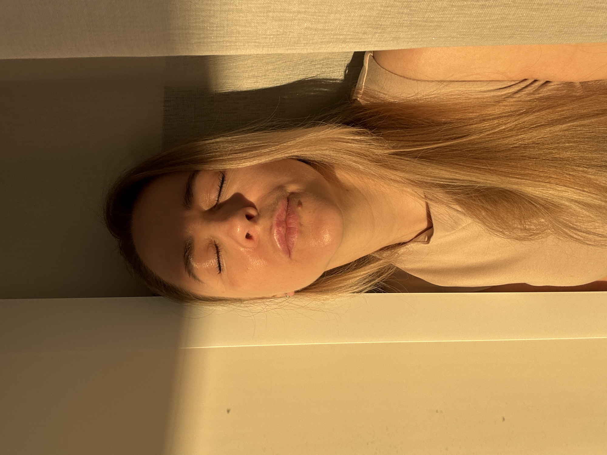 A woman with long blonde hair is lying down, with her eyes closed and her face illuminated by sunlight. She is wearing a light beige t-shirt. The background consists of a light beige fabric above her and a white, possibly wooden, surface below her. The location cannot be determined from the image; Chaikovsky, Russia cannot be determined.
