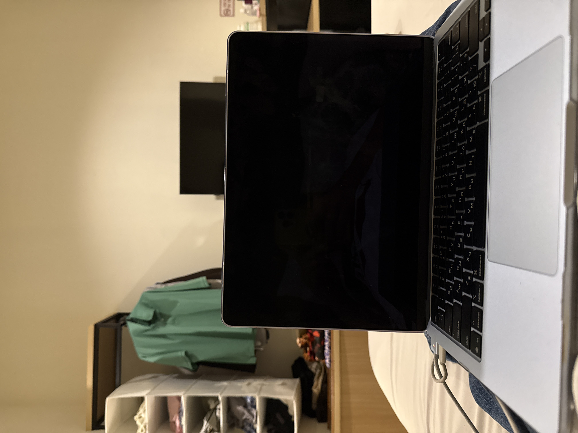 The image shows a close-up view of a laptop and a tablet placed on what appears to be a bed. The laptop's keyboard is visible, and the tablet's screen is black. In the background, a wall with a TV mounted on it can be seen. Beneath the TV, there's a clothing rack with a green garment and a white storage unit with compartments. Due to lack of geographical location details, I am unable to infer the location in North Kuta, Indonesia.
