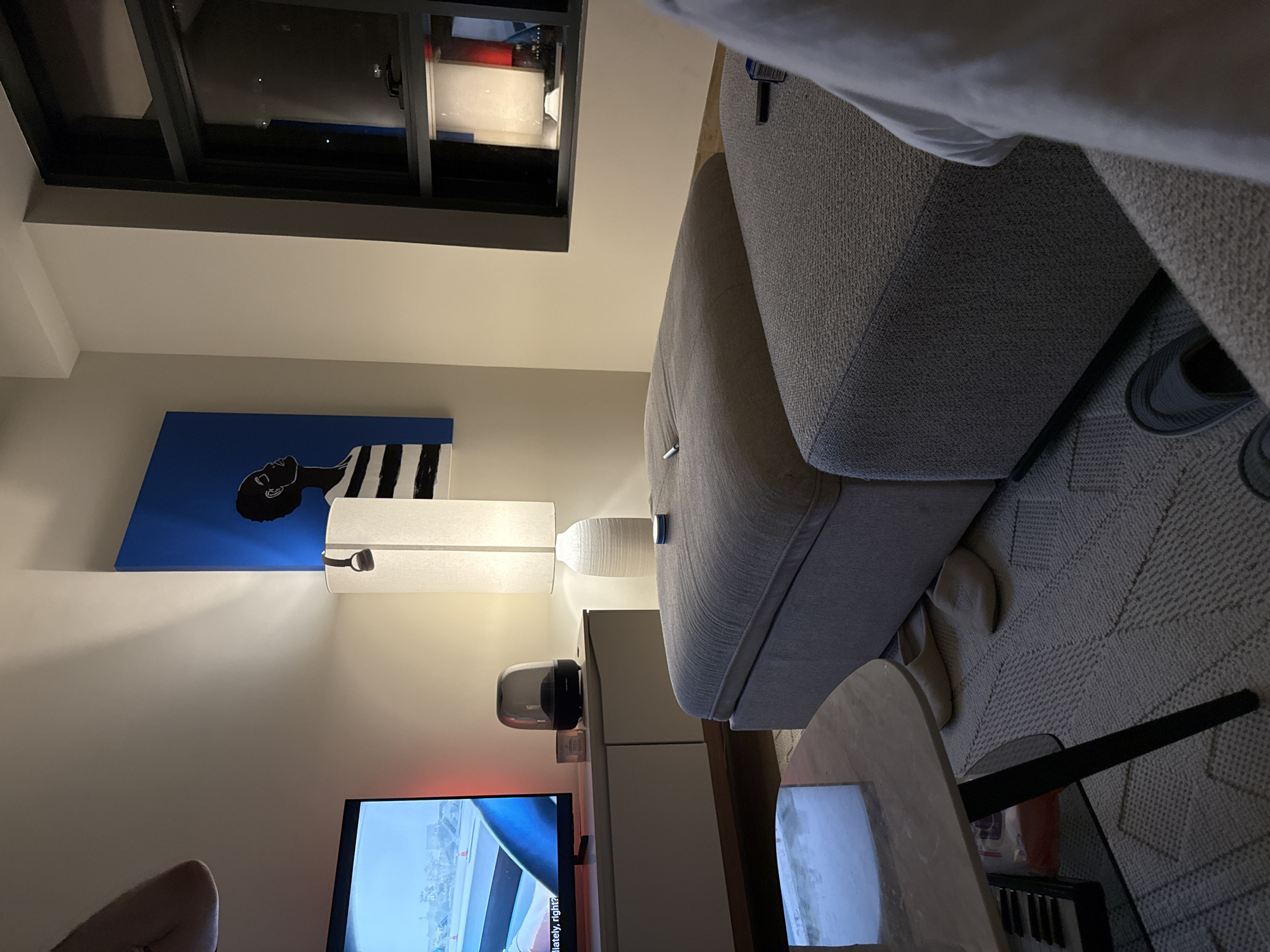 The image depicts a contemporary living room scene, likely within a residential apartment in Dubai, United Arab Emirates. The orientation of the image is rotated, with the ceiling appearing on the left and the floor on the right.
Dominating the scene is a large, plush grey sofa, positioned in the center-right of the frame. A portion of a person's arm and shoulder is visible on the far left, suggesting they are either seated or lying down in the room, partially obscured. In the foreground, a small, oval-shaped coffee table with a reflective surface is present. Beneath the coffee table, a small keyboard or synthesizer is visible.
On the wall to the left, a striking piece of modern art hangs. It features a black and white striped pattern behind the head and shoulders of a person rendered in silhouette against a vibrant blue background. Below the artwork, a tall, white rectangular lamp illuminates the wall with a soft glow. To the right of this lamp, a sleek, modern cabinet unit is visible, topped with a decorative vase and a dark, dome-shaped object, possibly a speaker or diffuser.
Further to the right, a television screen displays an image, with subtle LED lighting glowing from behind it. A small, round, dark-colored object is attached to the side of the sofa. A patterned rug covers the floor area beneath the sofa and coffee table.
The lighting suggests an indoor evening or nighttime setting, with artificial lights creating a warm and inviting ambiance. The overall aesthetic is modern and minimalist, indicative of a well-decorated urban living space. No text is explicitly legible within the image.