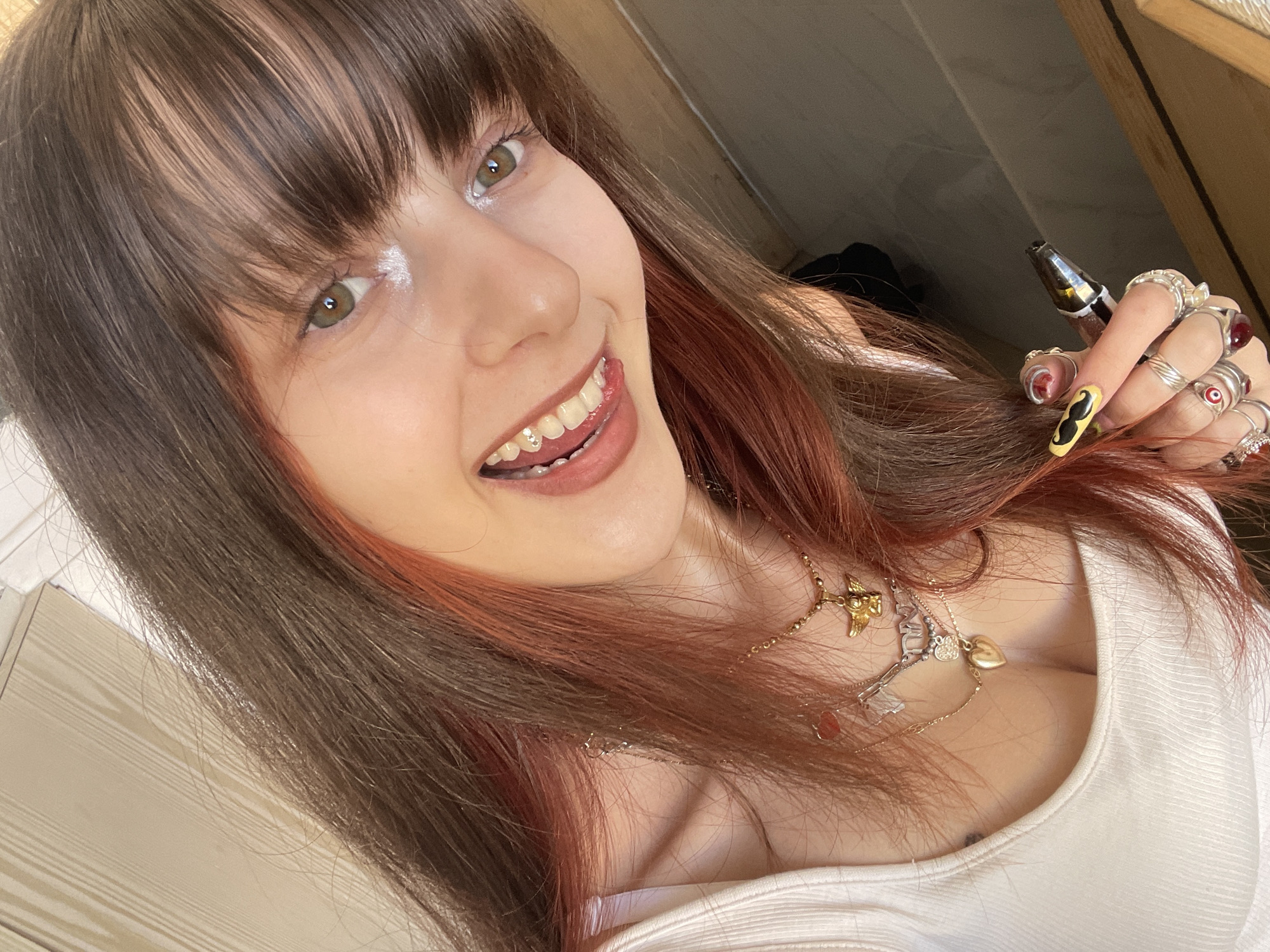 A close-up selfie shows a young woman, likely in Petah Tikva, Israel, smiling playfully and looking directly at the camera. She has long, dark brown hair with reddish-brown highlights or an ombre effect, and bangs covering her forehead. Her green or hazel eyes are bright, and a noticeable highlight emphasizes her cheekbone near her left eye. Her open-mouthed smile reveals small, sparkling jewels affixed to one of her lower front teeth, and her tongue is slightly extended.
She is wearing a white, ribbed top with a scoop neck and multiple gold-toned necklaces, including one with a cherub or angel pendant and another with a heart charm. Her left hand, held near her face, is adorned with numerous rings, some featuring stones like a red gem and an evil eye motif. Her long, manicured nails are painted, with one prominent nail showcasing a black mustache design on a yellow background. She holds a small, dark object, resembling a vape pen, between her fingers.
The background is softly blurred, indicating an indoor setting with light-colored surfaces, possibly walls or wooden panels, suggesting a home environment. The woman's expression conveys happiness and playfulness. No visible text is present in the image.