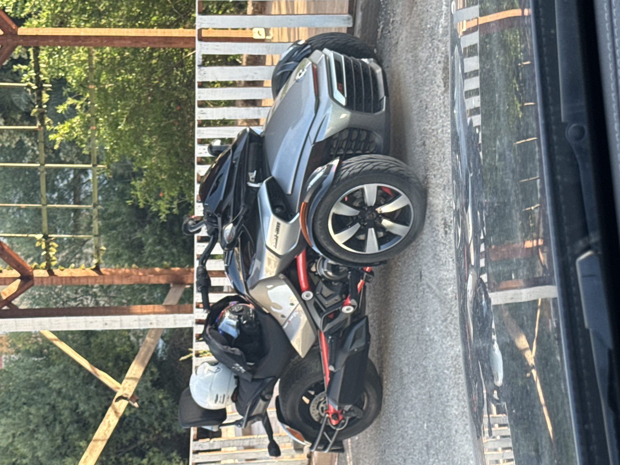 The image shows a silver Can-Am Spyder F3-S parked on a gravel surface. The three-wheeled vehicle is positioned in front of a white wooden fence and a rustic wooden canopy structure. A white helmet and a black backpack are visible on the rear seat of the vehicle. The background consists of lush green trees. The scene appears to be outdoors, possibly in a residential or recreational area. Based on the typical architecture and vegetation, this location could be in the city of Budva, Montenegro, a coastal city known for its scenic beauty and outdoor spaces.