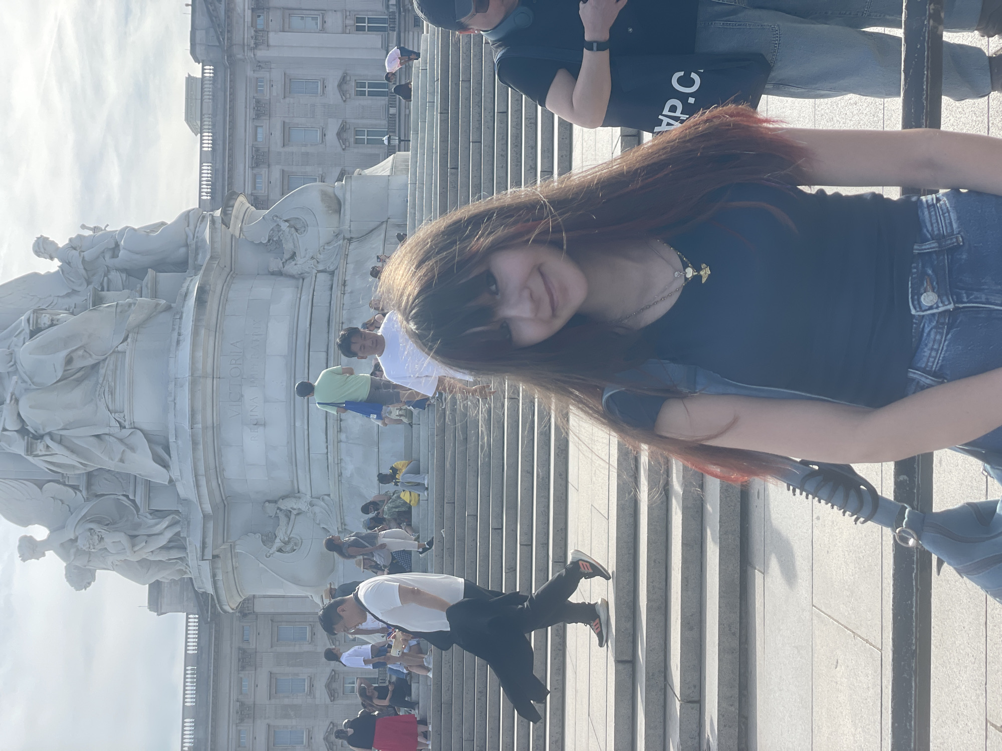 The image depicts the Victoria Memorial in front of Buckingham Palace, London. In the foreground, a young woman with long, reddish-brown hair and bangs is looking at the camera, smiling. She is wearing a black sleeveless top and blue denim jeans, with a gold necklace featuring a heart and wings pendant. A blue bag is slung over her left shoulder. To her right, a person wearing a black t-shirt, black baseball cap, and sunglasses is visible from the chest down, holding a black bag with white lettering that reads "YP.C.".
In the mid-ground, numerous people are gathered on and around the steps leading up to the monument. A man in a white t-shirt and black pants is walking down the steps, holding a black jacket over his arm. Other individuals, dressed in casual attire, are seated or standing on the steps, engaging in various activities. The background features the grand architecture of Buckingham Palace. The sky is bright and overcast.