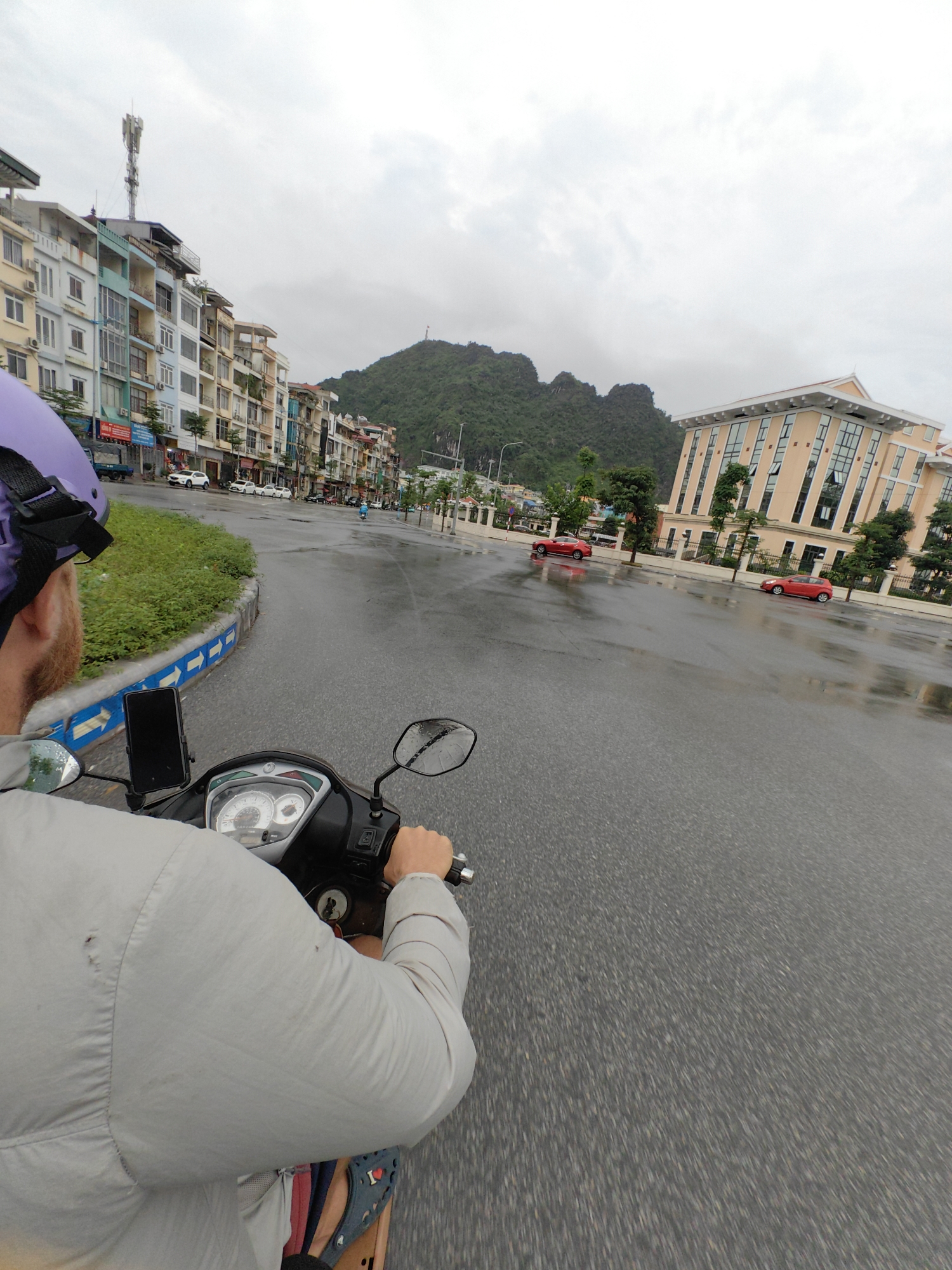 A first-person perspective captures a person riding a scooter on a wet road in Hong Gai Ward, Vietnam. The rider wears a purple helmet and a light gray jacket. A smartphone is mounted on the scooter's handlebars. The road surface is visibly wet, reflecting the overcast sky. Buildings line the left side of the road, while a large, light-colored building stands to the right. In the distance, a mountain range rises, and a red car is seen driving on the road. Other parked cars are on the side of the road.