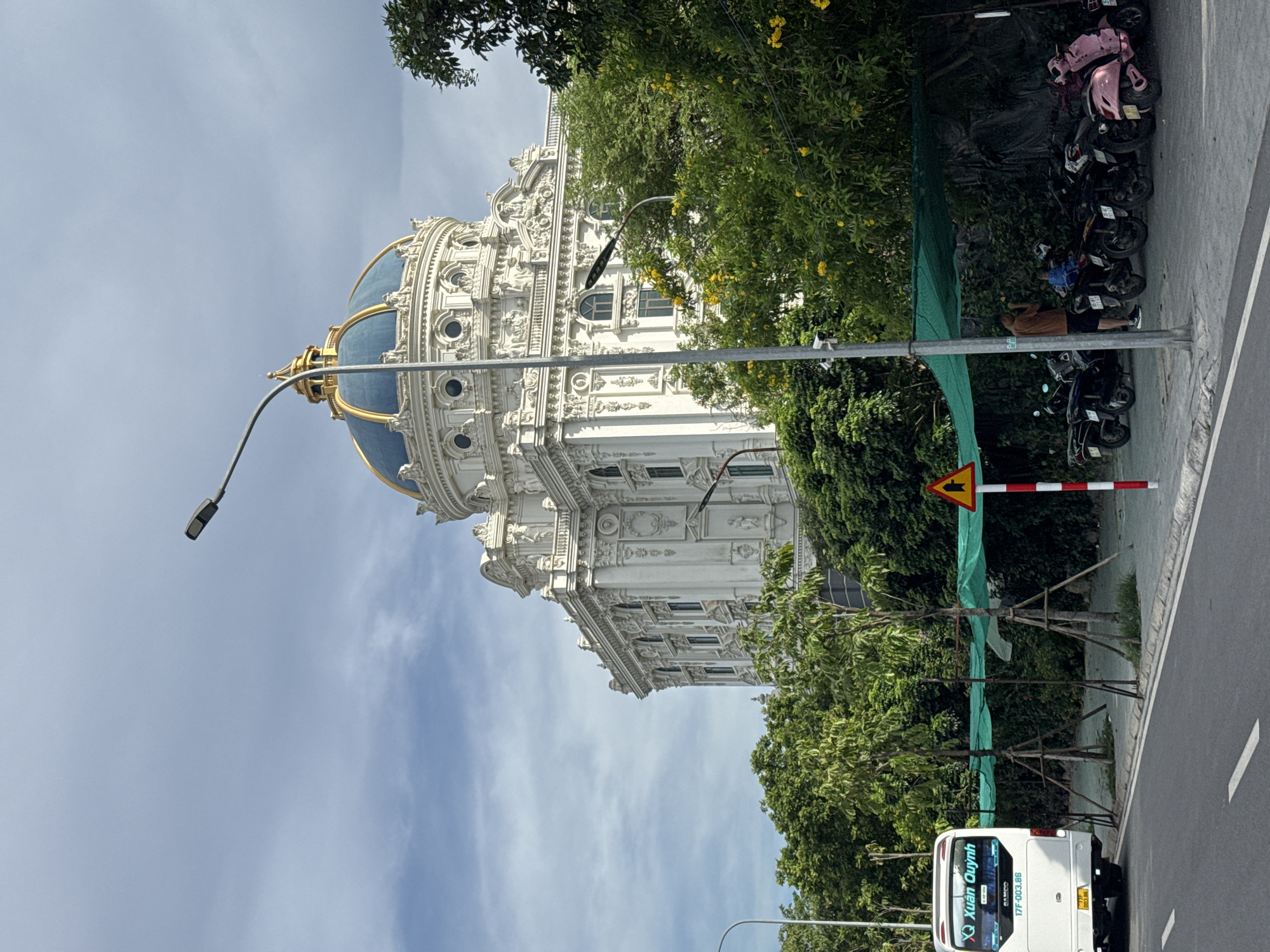 The image shows a large, ornate white building with a blue and gold dome, located in Hoa Lư Ward, Vietnam. In front of the building, there is a street with a white bus on the left side. Further down the street, parked motorcycles are visible under a green awning. Trees with green foliage and yellow flowers line the area between the street and the building. A street light stands on the right side of the image. A person is standing near the parked motorcycles. The sky is partly cloudy.