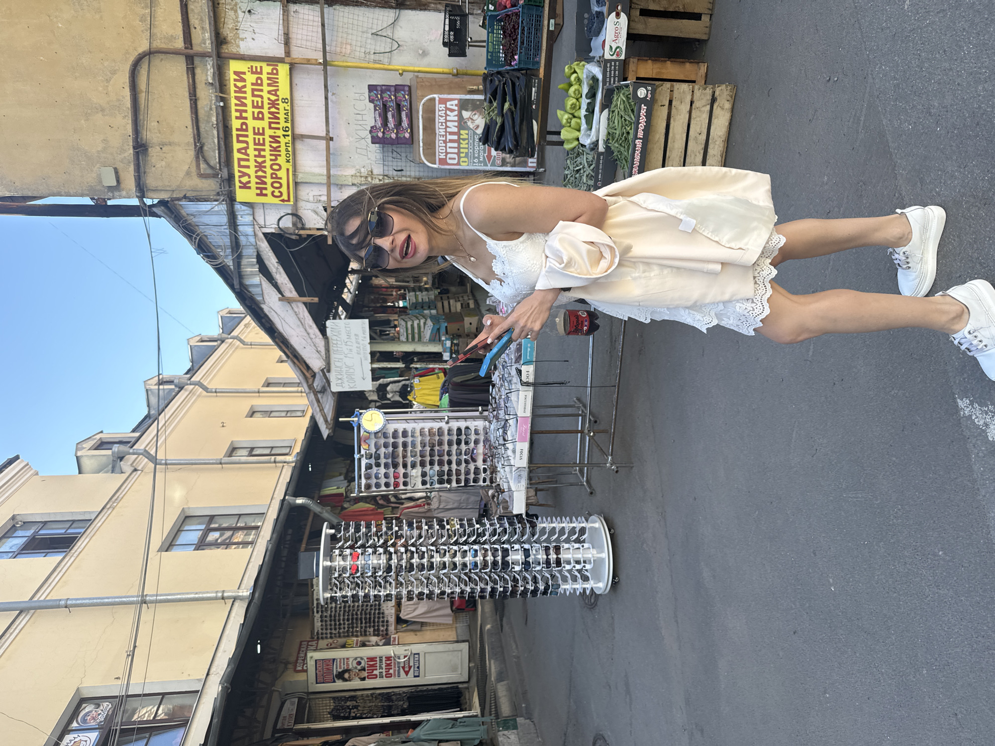 A woman in a white lace dress and white sneakers stands in an outdoor market, holding two mobile phones. She is wearing large sunglasses and a pale shawl draped over her shoulders. Behind her, various market stalls are visible, including displays of sunglasses, clothing, and fresh produce such as eggplants, green beans, and cherries. Signs in Russian indicate the types of goods sold, such as "Купальники. Нижнее белье. Сорочки. Пижамы" (Swimwear. Underwear. Shirts. Pajamas) and "Корейская оптика" (Korean optics). The architecture suggests an older urban setting, consistent with markets found in Saint Petersburg, Russia. The lighting indicates it is daytime with clear skies.