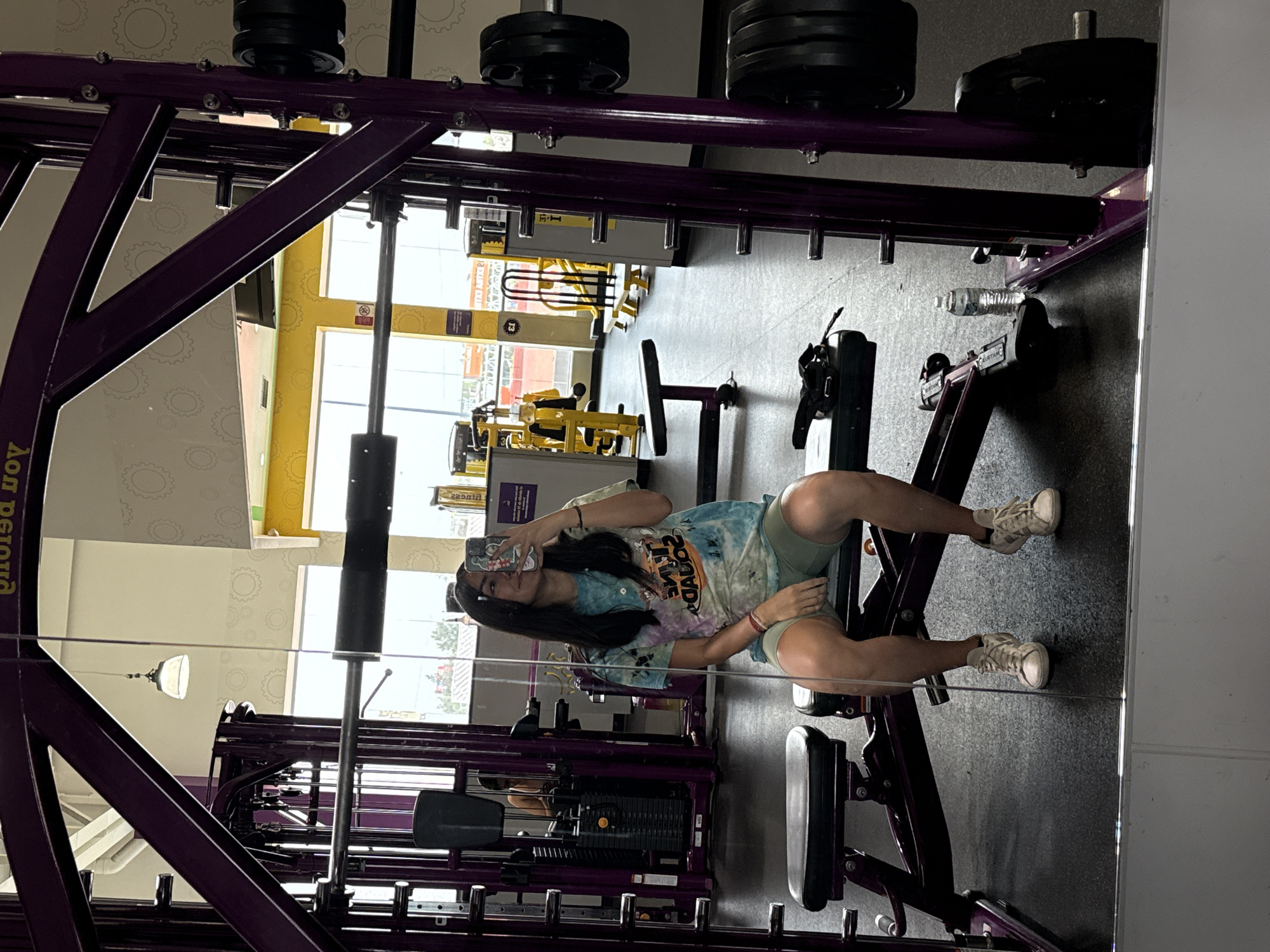 A young woman sits on a leg extension machine in a Planet Fitness gym, presumably in Puebla, Mexico. She is taking a selfie, holding her phone in her left hand. She is wearing a tie-dye t-shirt and green shorts, and white athletic shoes. The gym equipment is purple. In the background, other gym equipment is visible and a large window showing the exterior of the building.