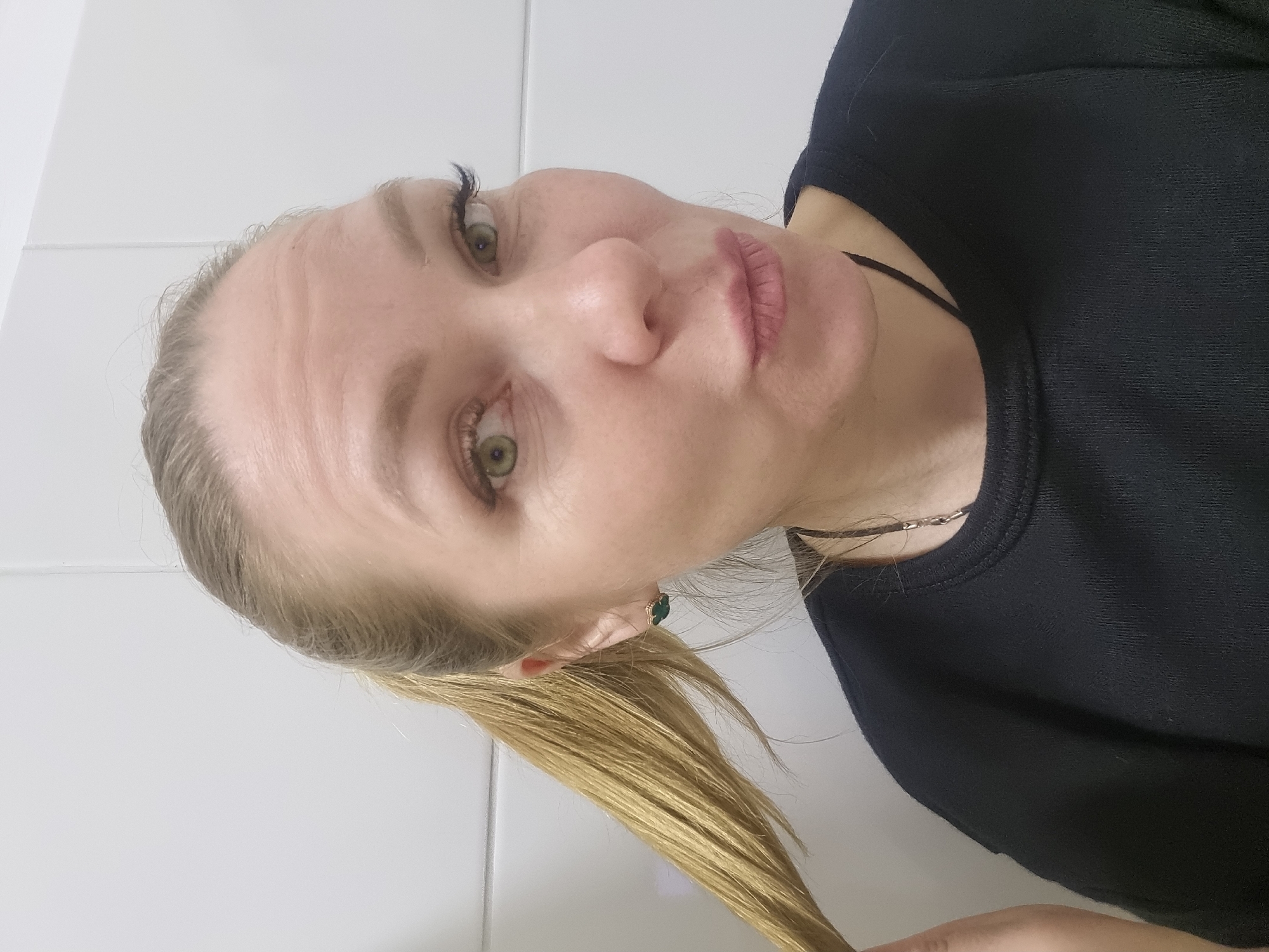 A woman with blonde hair pulled back in a ponytail is positioned against a white tiled background. She is wearing a black top and a necklace. She appears to be holding a strand of her hair. Her skin tone is fair. She has green eyes and dark eyelashes. The photo is taken from a close-up angle, slightly angled towards her face. The location is not discernable from the image.