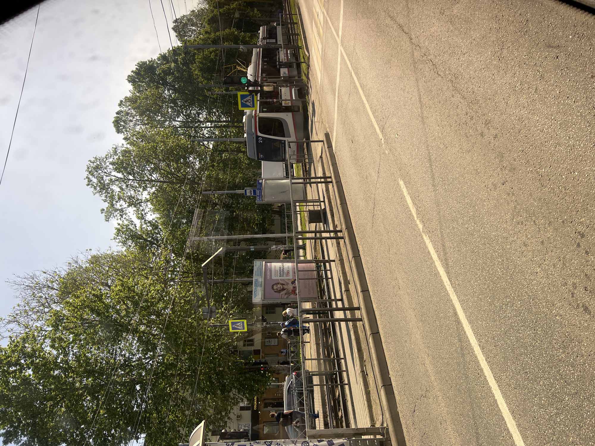The image shows a street scene in Krasnodar, Russia. There is a road with a white line dividing lanes and a tram line with trams parked along it. There are trees lining the street and pedestrian crossing signs visible. People are waiting at the tram stop, indicated by a bus stop sign. There is a poster or advertisement at the tram stop.