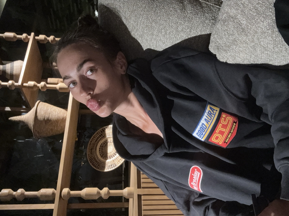 A woman wearing a black hoodie lies against a gray surface. The hoodie has patches with the words "GOOD AURA" and "OTS RACING GOODS." Behind her is a shelf-like structure with woven baskets on it. She is making a kissing face towards the camera. The lighting is dark, casting shadows on the subject and the background.