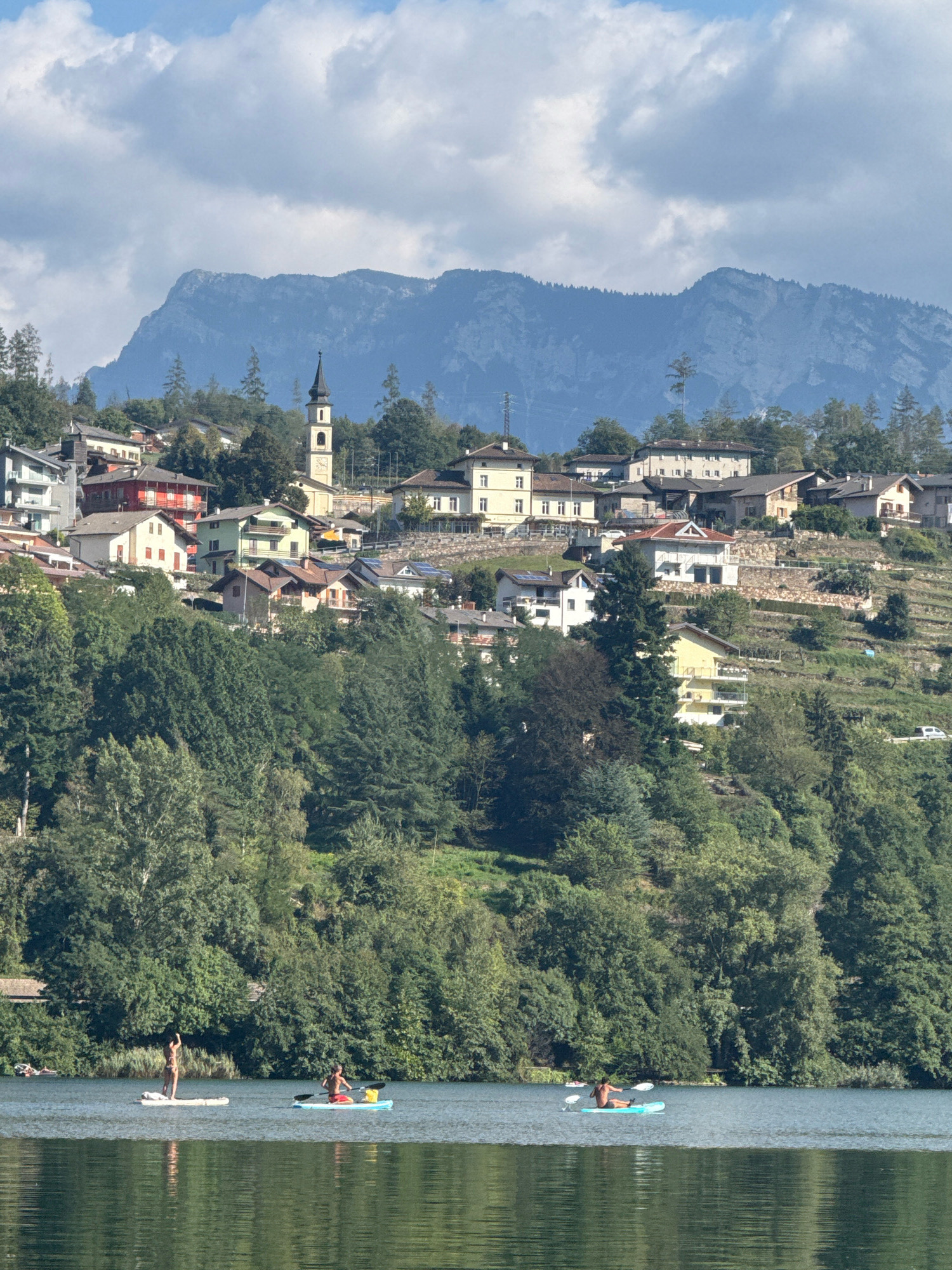 This is a landscape photograph featuring a serene lake in the foreground with a town nestled on a hillside in the background, beneath a mountainous terrain. The lake's surface is calm, reflecting the sky and the surrounding greenery. On the water, three individuals are engaged in water activities. In the left mid-ground, a person stands on a paddleboard. To the right of this person, another individual is seated on a paddleboard, holding a paddle. Further to the right, a third person is also seated on a paddleboard, paddling.
The hillside town is characterized by a dense cluster of buildings, varying in architectural style and color. Prominently visible is a church with a tall, slender spire. The buildings are interspersed with lush green trees and vegetation, with some terraced areas suggesting cultivation. Beyond the town and trees, a rugged mountain range forms the backdrop, its peaks partially obscured by a light haze or clouds. The sky above is a mix of blue and white clouds. The overall scene depicts a tranquil and picturesque view of a lakeside town in a mountainous region. Based on the context of Pergine Valsugana, Italy, this scene likely depicts Lake Caldonazzo or Lake Tenna, with the town being Pergine itself or a smaller village within the Valsugana valley.