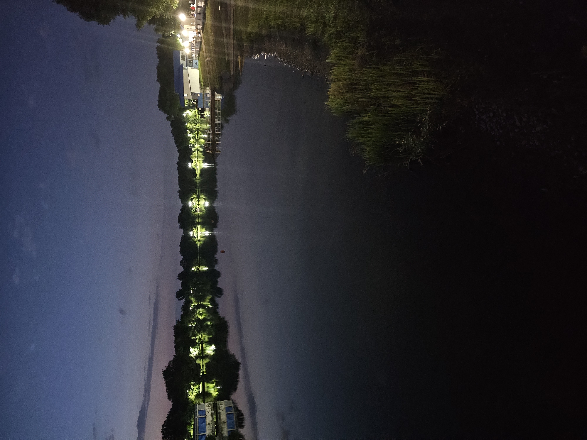 This is a nighttime photograph of a lake with a tree-lined shore and a building on the right. The sky is a deep twilight blue with some scattered clouds. Lights from the building and along the shore reflect on the dark water. There appear to be people dining or socializing on the patio of the building on the right. The image is of Lake Itkul in Cherkessk, Russia.