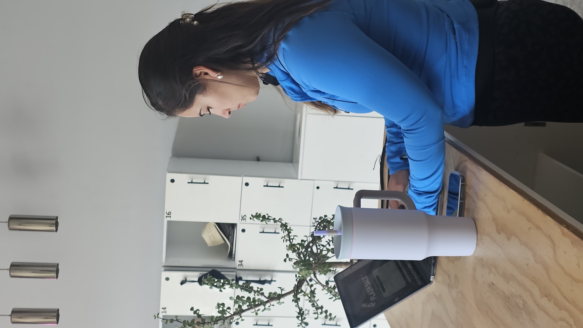 A woman with long, dark hair, wearing a blue long-sleeved shirt and black pants, is working at a wooden desk. She is looking down at a phone resting on the desk. A white, handled tumbler with a straw stands beside the phone. A tablet with the words "FLASH SALE" on its screen is visible on the desk, partially obscured by a potted plant. In the background are white lockers with black handles. The numbers "16", "34", and "35" are visible on the lockers. Above the lockers, pendant lights hang from the ceiling. It is not possible to determine the location as Puebla, Mexico, based on the provided image.