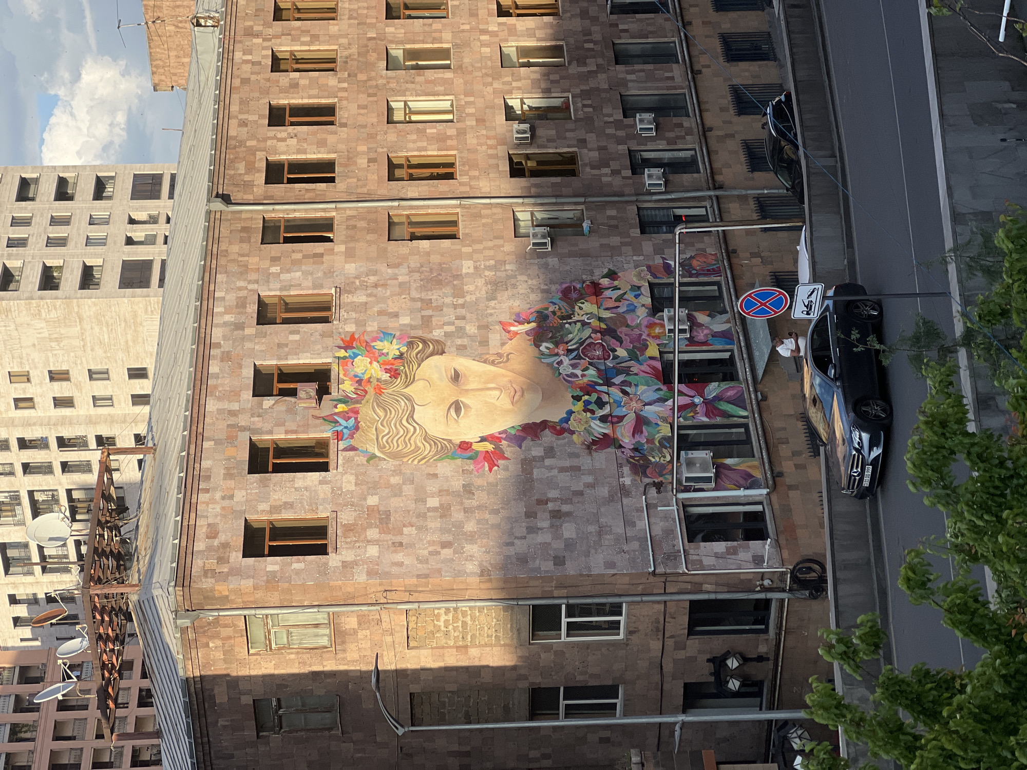 The image captures a view of a building in Yerevan, Armenia, featuring a large mural of a woman's face adorned with colorful flowers and leaves. The building itself is constructed of light brown stone bricks and has numerous windows, some of which have air conditioning units installed. A street runs alongside the building, where a black Mercedes sedan is parked. A "no parking" sign is visible near the vehicle, suggesting parking regulations in the area. Part of another tall building is seen on the left side of the image. Green foliage from a tree is on the bottom right corner of the shot, adding a touch of nature to the urban scene. Additionally, there's a collection of satellite dishes mounted on a pole attached to another building.