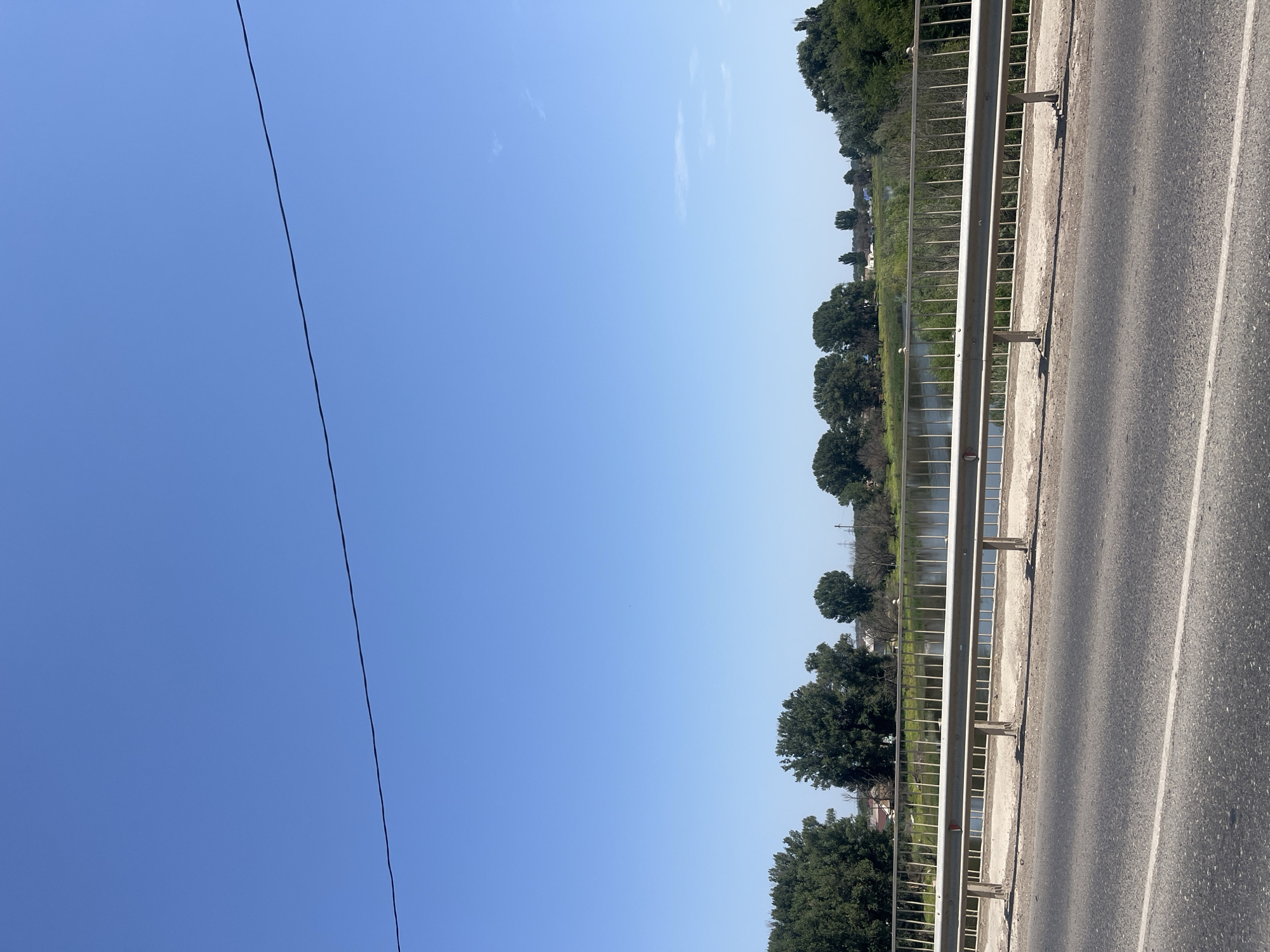 The image captures a roadside view of a calm body of water with a tree-lined bank. A metal guardrail separates the asphalt road from a paved pathway, which overlooks the water. In the distance, several houses and buildings are visible amidst the trees. The sky is a clear, bright blue, with a single black wire running horizontally across the top portion of the frame. The scene appears to be located in Kamyzyak, Russia, as indicated by the presence of typical rural architecture and vegetation common to the region.