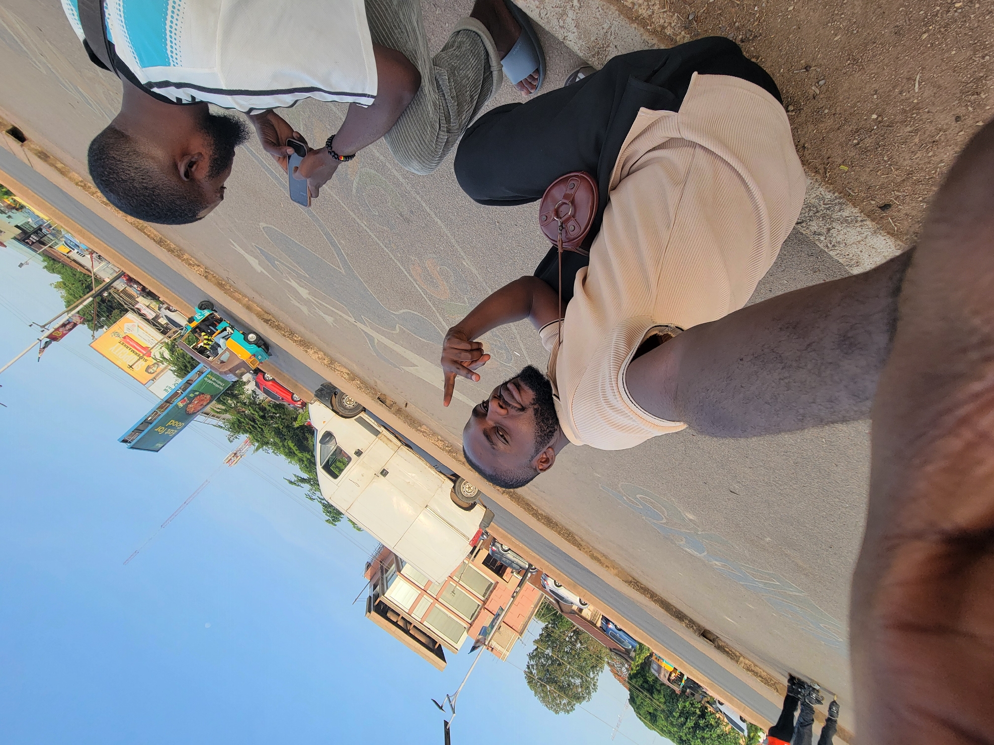This image depicts a casual outdoor scene in what appears to be an urban or semi-urban setting in Jos, Nigeria, under bright, clear daylight. The photograph is taken from a low angle, possibly a selfie.
In the foreground, two men are visible. On the right, partially visible, is a man with a dark beard, wearing a light-colored (beige or tan) ribbed shirt. He is pointing his right index finger towards the other man. A small, round, brown leather bag with a short strap hangs from his shoulder or chest.
To his left, another man with a dark beard is engrossed with a smartphone held in both hands. He wears a white and light blue patterned shirt with black trim, over a dark undershirt, and light-colored (possibly olive green or khaki) ribbed pants. A beaded bracelet is on his left wrist. Adjacent to him, a third person's feet are visible, wearing light blue slides.
The men are situated on a dirt or gravel shoulder next to a paved road. In the background, a white delivery van is parked on the side of the street. Further down the road, a colorful Keke Napep (motorized tricycle, common in Nigeria) is visible, along with a red car and other vehicles. The road surface has some faded markings.
The background also features several buildings, including a multi-story structure with reddish-brown and white elements. Multiple billboards are present; one yellow sign clearly reads "SUPERMARKET," while a larger blue billboard, visible upside down, shows the text "good for eat" (or "eat for good"). The sky is clear and blue, indicating a sunny day. Green trees and streetlights are scattered throughout the scene. The overall mood is relaxed and informal, capturing a candid moment.