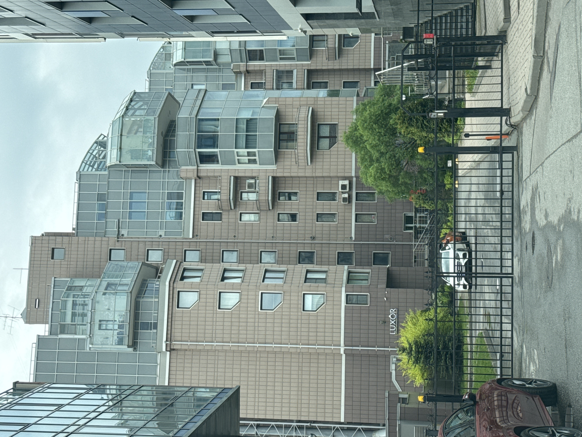 The image shows a multi-story building in Saint Petersburg, Russia, identified by the "LUXOR" sign on its facade. The building has a modern architectural style with numerous windows and glass extensions. A black metal fence runs along the sidewalk in front of the building. A red car is visible in the foreground, partially obscured by the fence. A white car is also visible through the fence, parked near the building. The environment appears to be a residential area, with other buildings visible in the background. The weather is overcast.