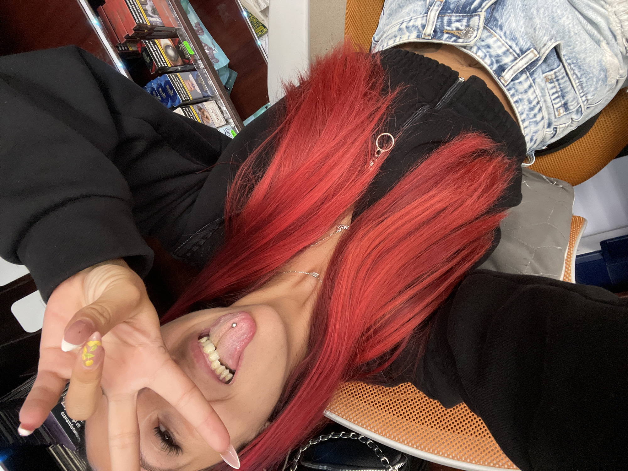 A young Caucasian female with vibrant red hair is taking a selfie. She is wearing a black zip-up hoodie and light-wash denim jeans. Her tongue is pierced with a silver barbell, and she is extending her right hand, forming a peace sign over her face. Her fingernails are long and painted with a French tip design adorned with small yellow flowers. The background appears to be an indoor environment, possibly a convenience store or kiosk, with shelves displaying packs of cigarettes. Based on the Bulgarian text visible on some of the cigarette packs, the location is likely Sofia, Bulgaria. The overall scene is casual and personal, captured in a selfie-style photograph.