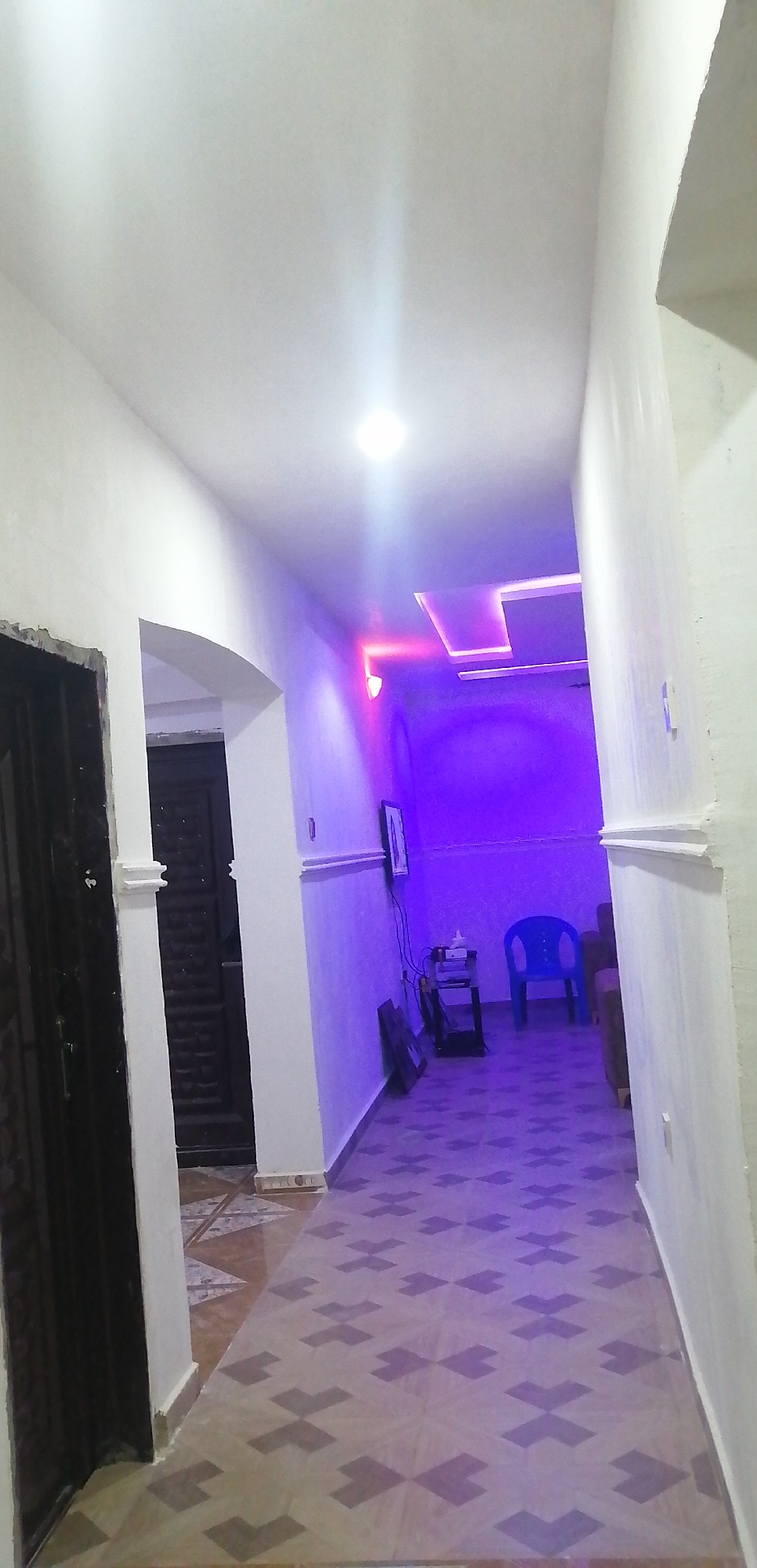 The image depicts an indoor hallway with modern lighting. The walls are painted white and have subtle decorative moldings. The ceiling is white and features a square recessed light fixture that is emitting a bright light. There is also a long, recessed lighting fixture on the ceiling further down the hallway, emitting purple and pink light. The floor is tiled with a patterned design in shades of beige and brown.
On the left side of the hallway, there is a dark-colored door with ornate carvings. Adjacent to it, a white pillar with decorative moldings supports an archway.
Further down the hallway, on the right side, there is a blue plastic chair. Behind it, a dark-colored sofa is partially visible. A black shelf unit is placed against the wall, holding a television, a small stack of items, and a framed picture. Another framed picture is leaning against the wall near the shelf. The walls are illuminated with ambient purple light, creating a striking visual effect. The overall impression is that of a contemporary and stylish interior space.