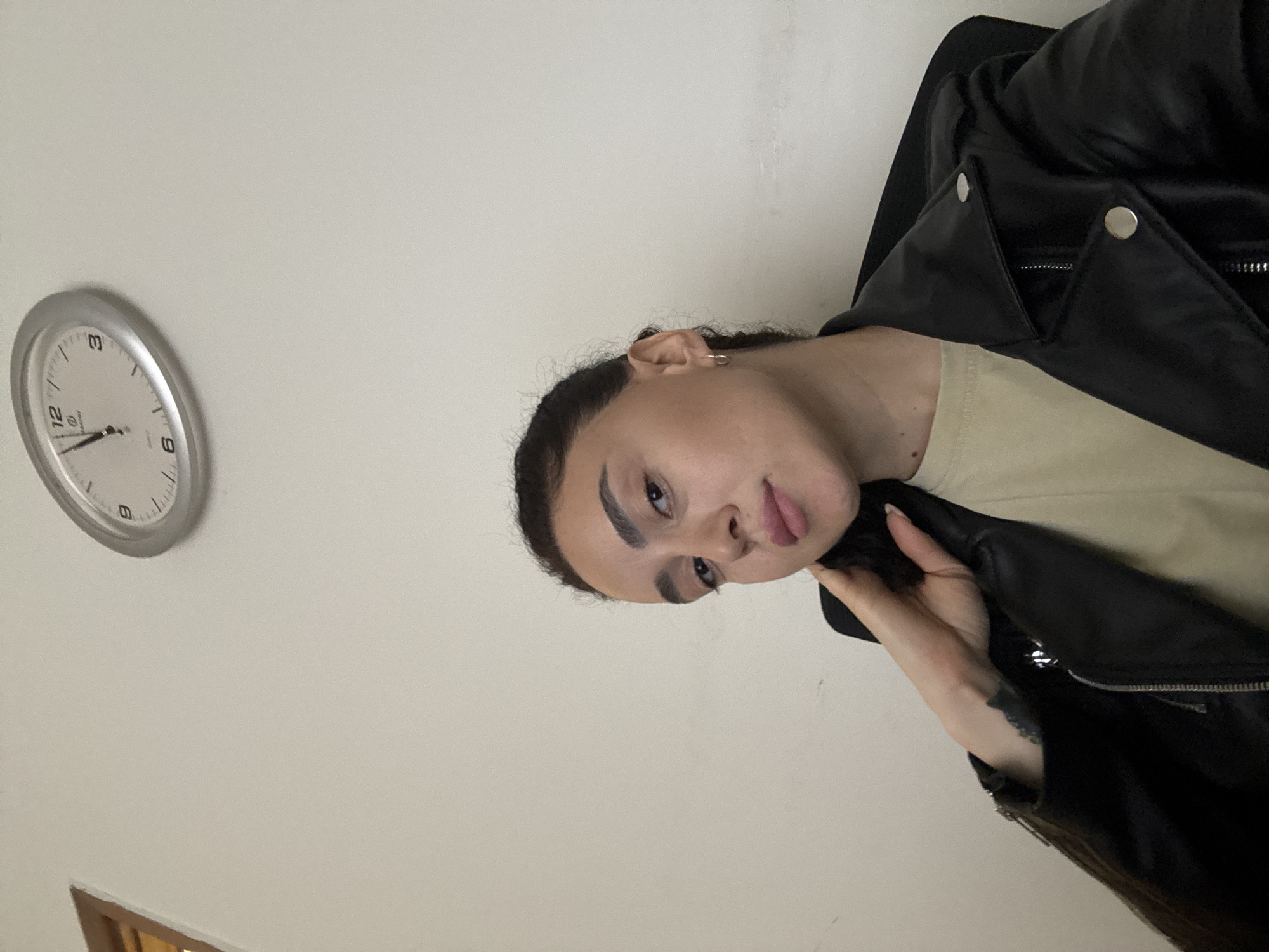 A person with long, dark hair and prominent eyebrows is wearing a black leather jacket and a light-colored shirt. They are holding the collar of the jacket with their left hand, which has a tattoo on the inner forearm. A silver analog clock is mounted on the wall behind them, reading approximately 1:50. A portion of a wooden doorframe is visible in the bottom left corner. The background appears to be a plain white wall. The location in the city of Tbilisi, Georgia cannot be determined from this image.