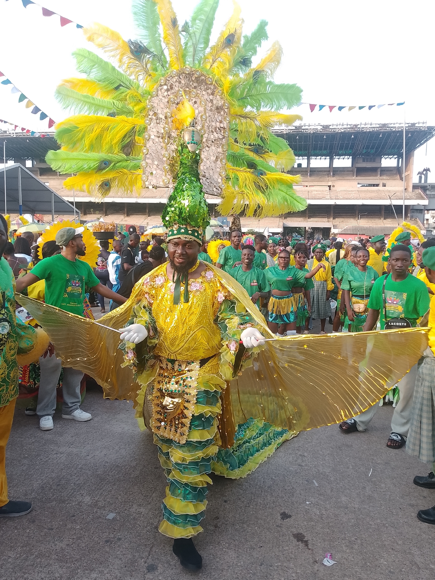A street scene in Lagos, Nigeria, shows a man dressed in a yellow and green peacock-themed costume standing on a paved road. The costume features a large feathered headdress, a sequined bodice, and wings held out to the sides. He wears white gloves and a sequined hat. A crowd of people, many wearing green shirts with "Lacoste" visible on one, surrounds him. In the background, a stadium-like structure is visible. Festive banners are strung above the crowd.