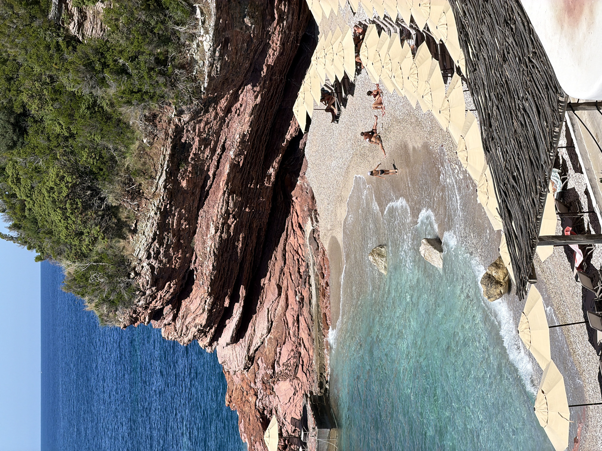 A beach scene is captured at Sveti Stefan, Montenegro. A rocky cliff face is on the left, descending to a beach with clear turquoise water. Several people are on the beach, some are sunbathing near the water's edge. Beach umbrellas and lounge chairs line the back of the beach. A barrier made of wooden sticks stands between the beach and a terrace. The sky is clear.