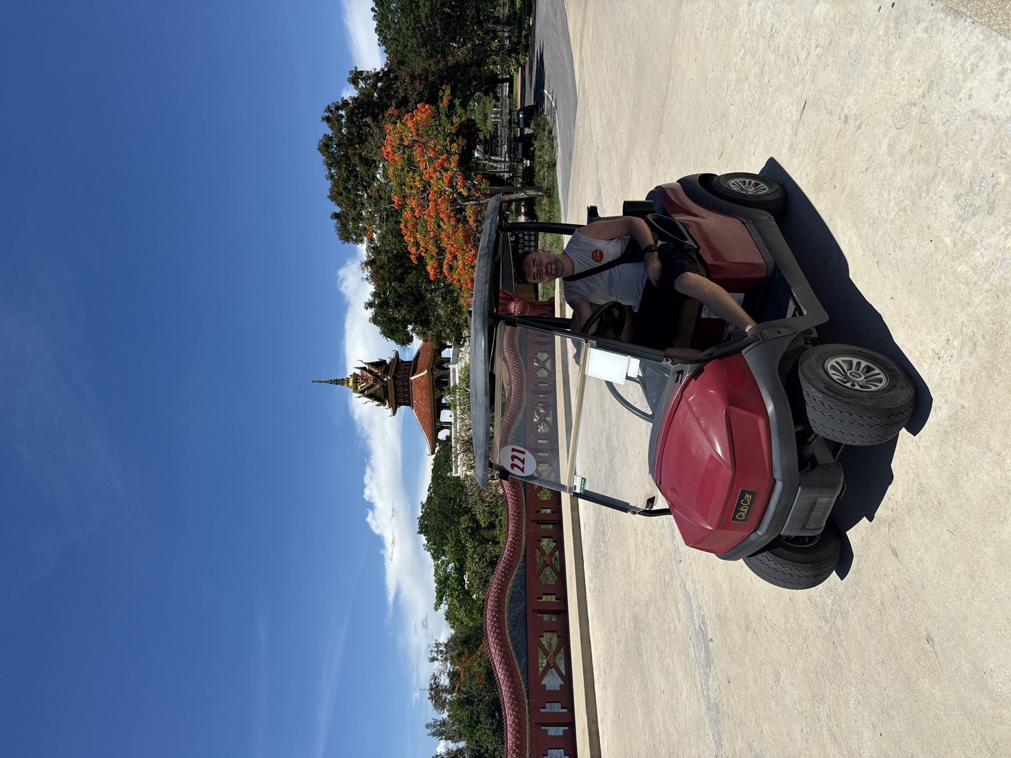 A man is sitting in a red golf cart on a concrete surface, with a building with ornate orange roofs and golden spire visible in the background. The number "221" is visible on the golf cart. A red fence with a dragon design is visible next to the cart. Trees and a clear blue sky are also visible. The location is Bang Pu Subdistrict Municipality, Thailand.