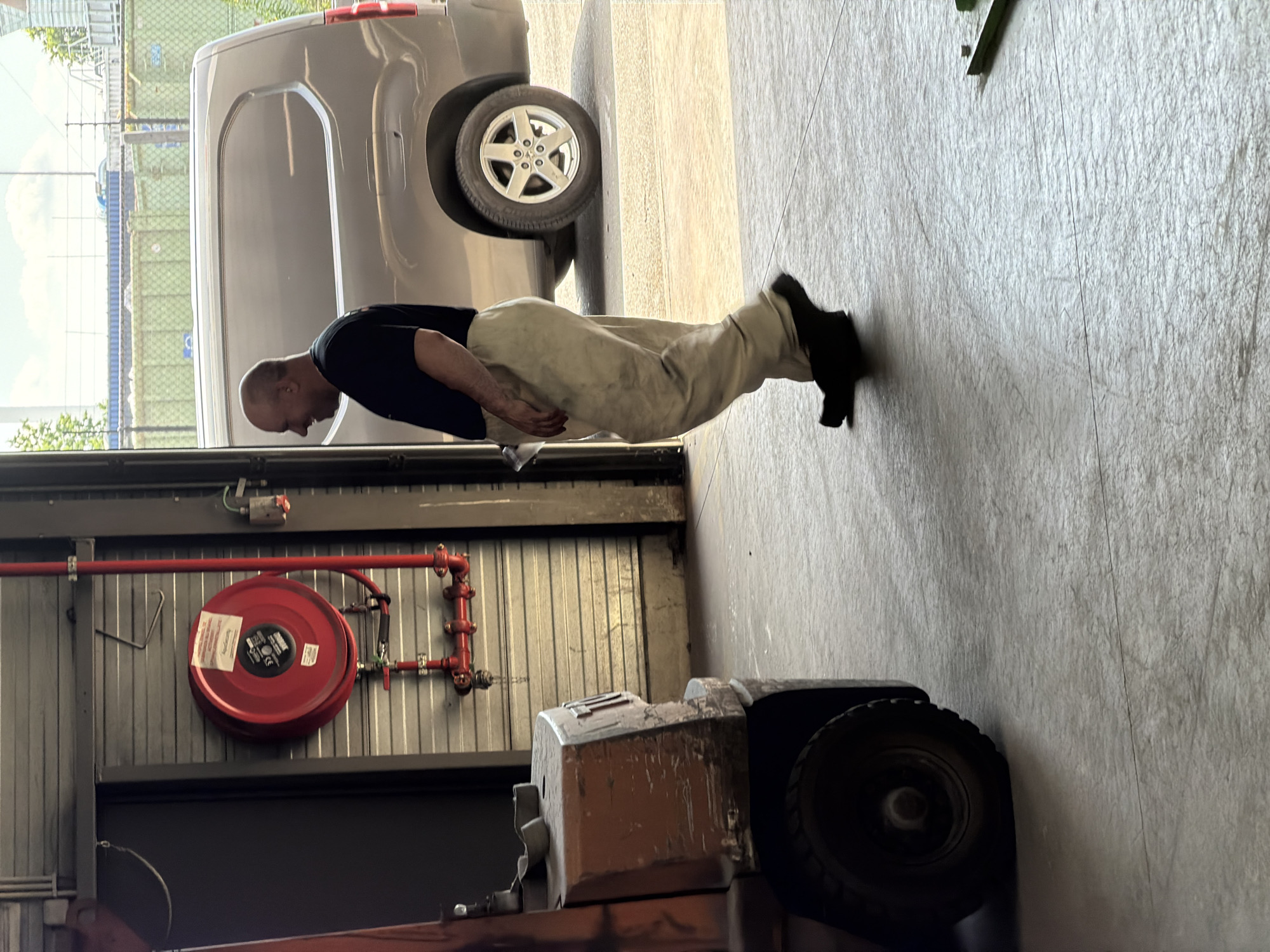 The image shows a man walking in front of a closed corrugated metal garage door. A red fire hose reel is mounted on the wall to the left of the door. A grey van is parked to the right of the garage door. A forklift is partially visible in the foreground to the left of the man. The man is wearing a dark t-shirt and light-colored work pants. He is bald and has a slight smile on his face. The scene appears to be an industrial or commercial setting. There is no identifiable information that places the image in the city of Mol, Belgium.