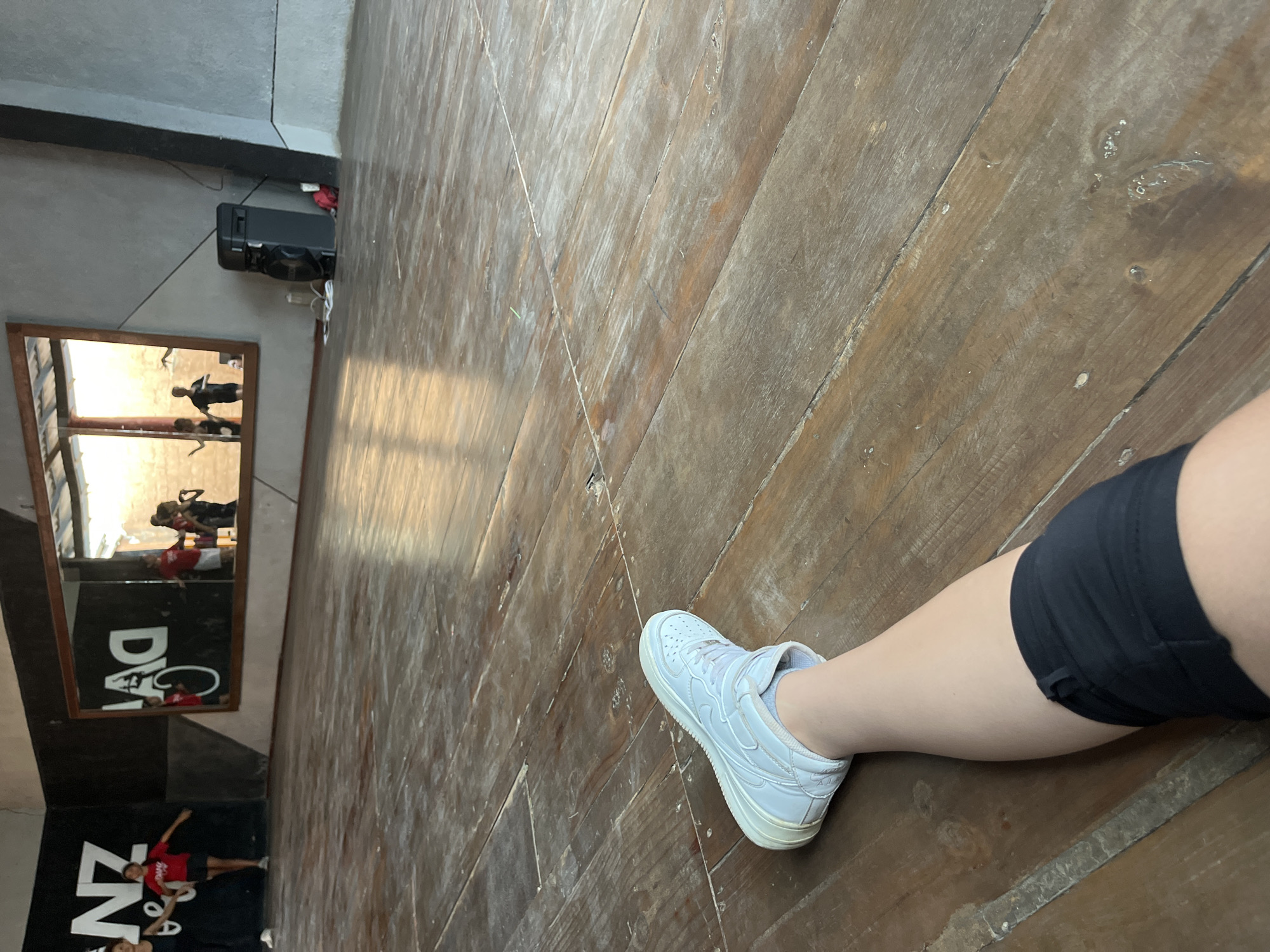 The image shows a person's leg and foot wearing a white sneaker on a wooden floor. The person is wearing black shorts. A mirror in the background reflects people engaged in dance or exercise. The location is a dance studio or gym with the logo "DZ" visible in the mirror's reflection. The city is not discernible from the image.