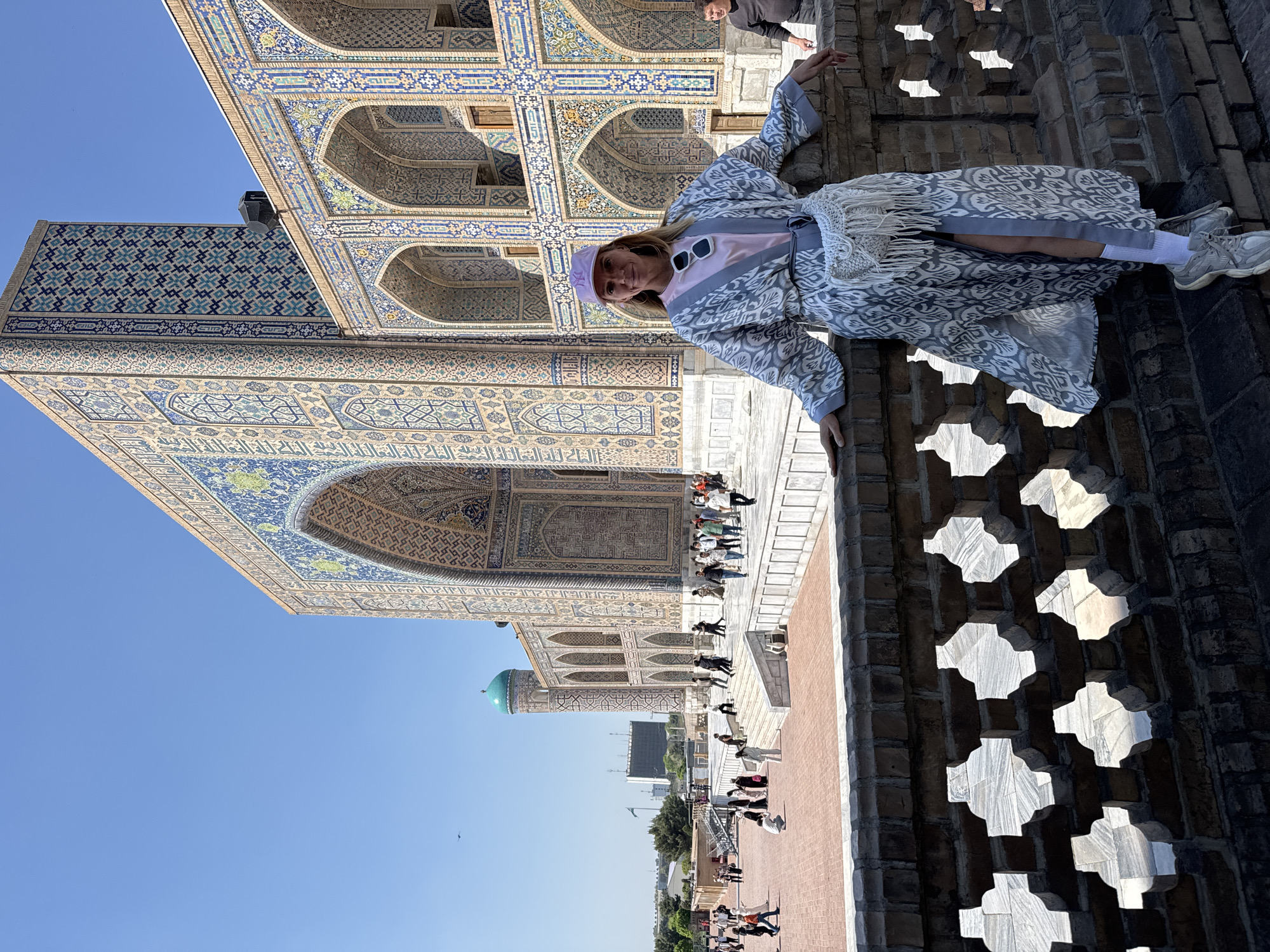 A medium shot depicts a person standing in front of the Tilya Kori Madrasah in Samarkand City, Uzbekistan. The person is wearing a long, patterned coat, a light pink top, white socks, and athletic shoes. They also have a light purple hat on. They are standing on a patterned brick structure.
Behind the person, the detailed mosaic work of the Tilya Kori Madrasah is visible, with arched openings and intricate patterns. In the background, a courtyard is partially visible with other people walking around. In the far background are city buildings under a clear blue sky.
