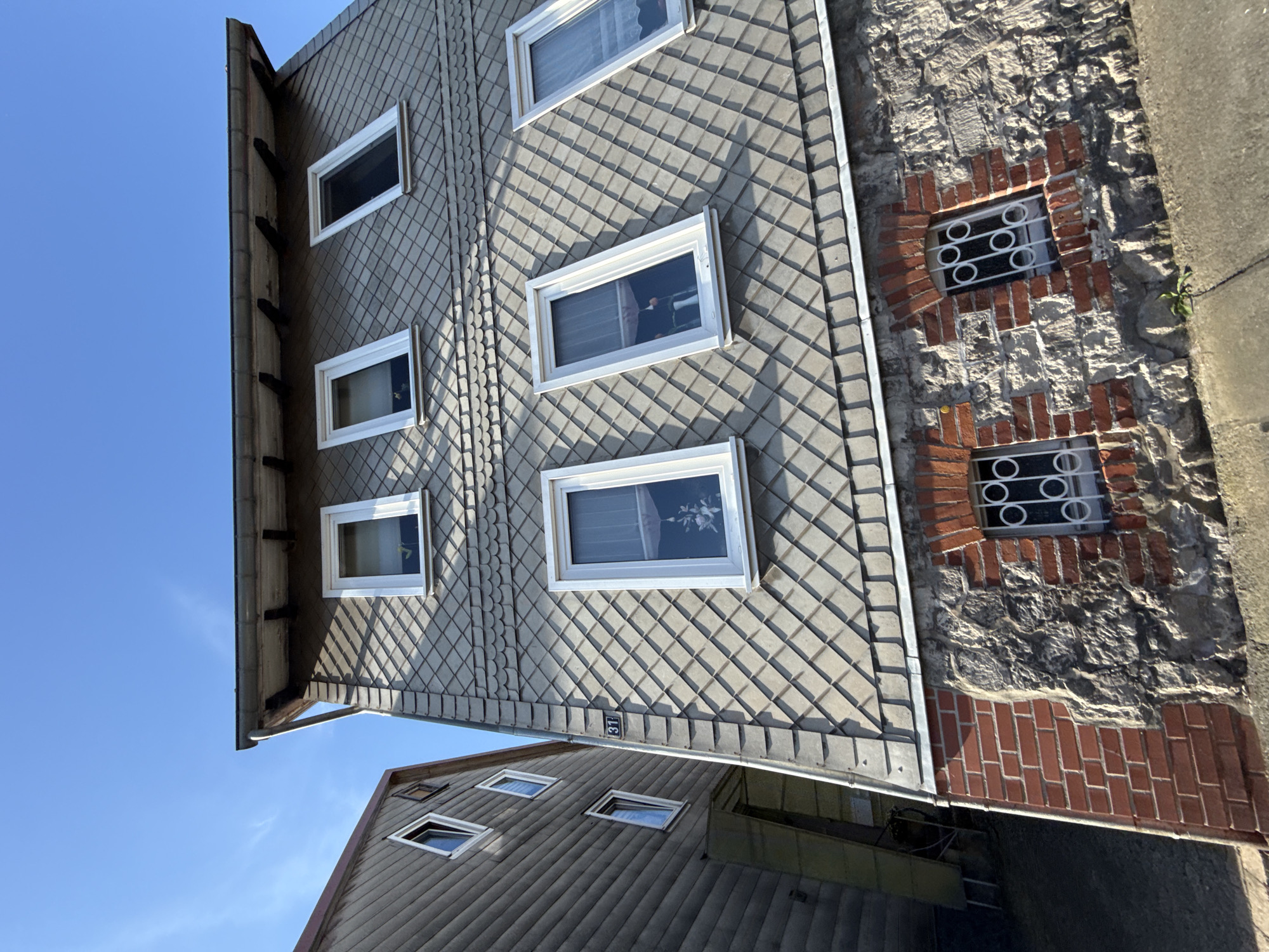 The image shows a low-angle view of two buildings under a clear blue sky. The building in the foreground has a facade covered in grey diamond-shaped shingles. The lower part of this building features a stone foundation with red brick accents around small, barred windows. The building to the left has a dark, horizontally paneled facade and appears to be slightly taller. The building in the foreground has a house number "31" displayed on a metal plate attached to the gutter. The scene is an exterior view of residential buildings.