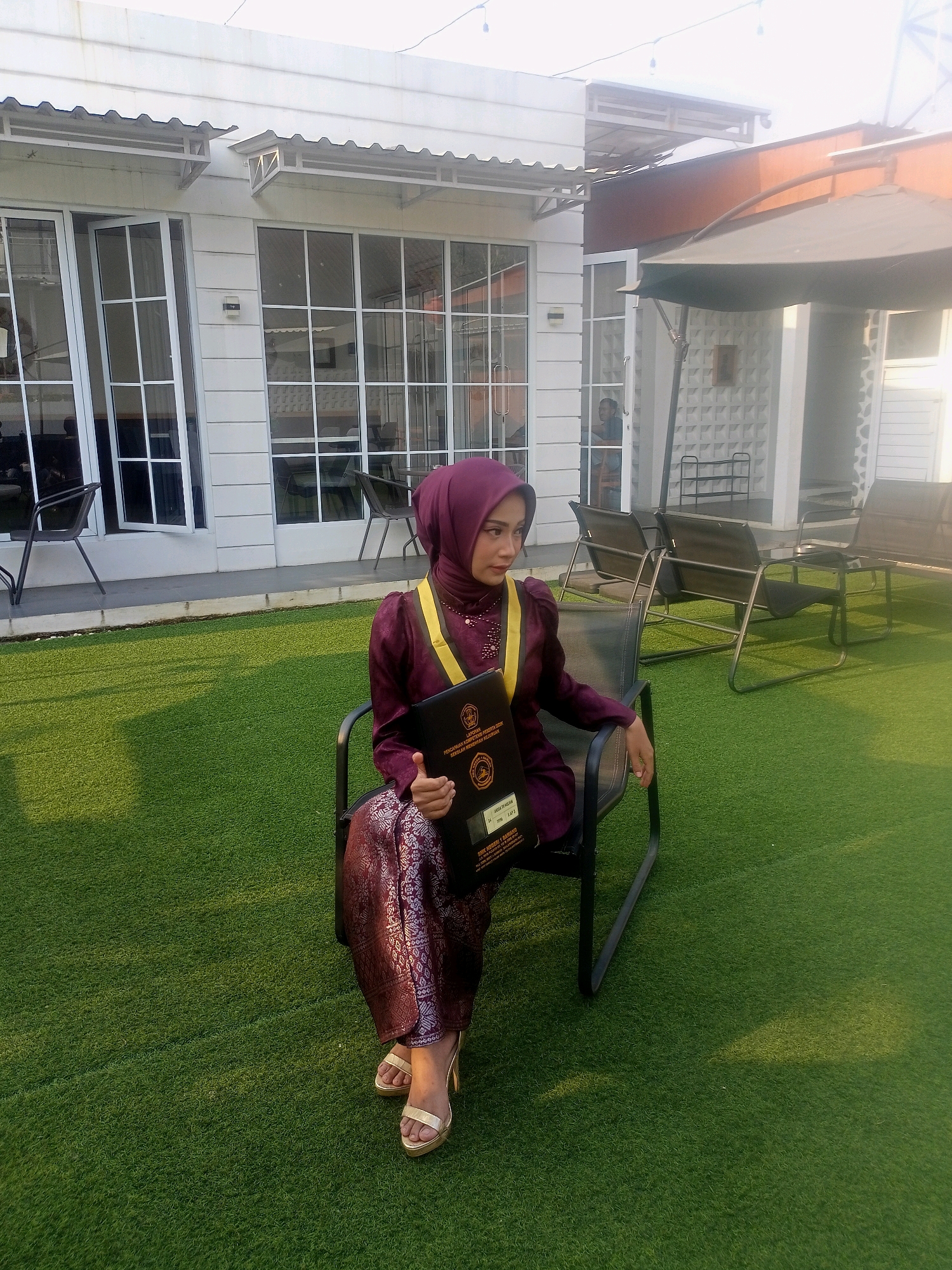 A young woman wearing a maroon hijab and a matching maroon graduation robe with a yellow sash is seated on a black metal chair on a patch of artificial turf. She is holding a black folder or certificate. The woman is wearing patterned maroon pants and gold high-heeled sandals. The background features a building with large glass windows and an outdoor seating area with chairs and umbrellas. The scene appears to be in an outdoor cafe or event space. The image does not contain any specific elements that would allow for precise identification of the location within Blambangan, Indonesia.