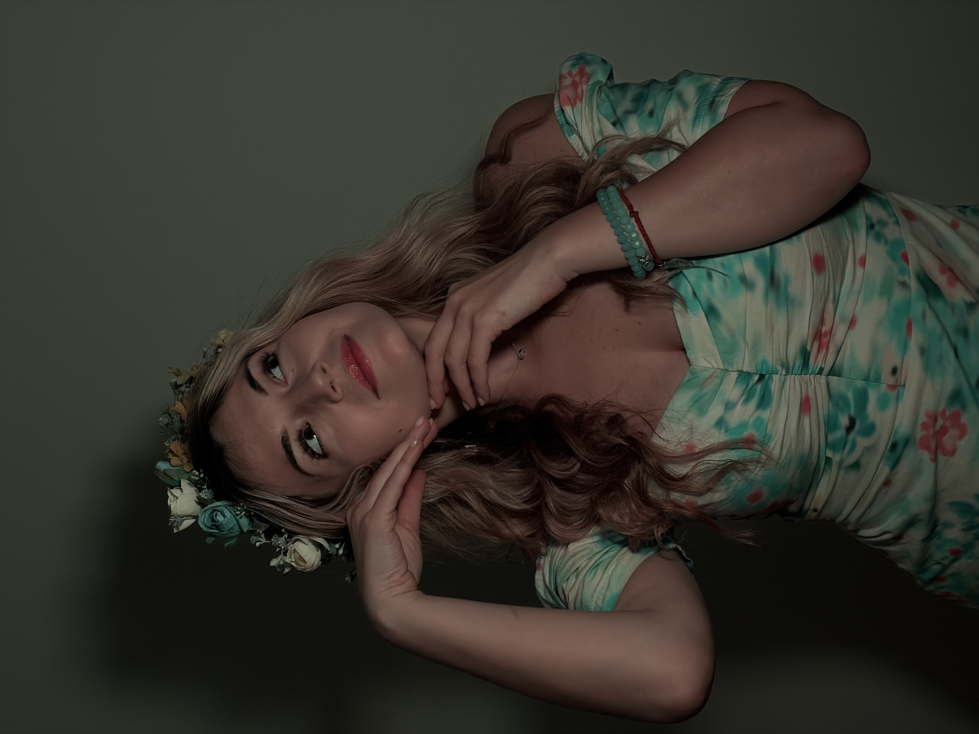 A young woman with long blonde hair wearing a floral crown and a light blue and pink floral dress poses against a dark, neutral background. She is wearing a blue beaded bracelet on her right wrist and is holding her hand near her neck and cheek. The image is oriented at an angle, with the woman tilted to the side. The lighting is dim, creating a moody atmosphere. There is no evidence to suggest that the picture was taken in Budva, Montenegro.