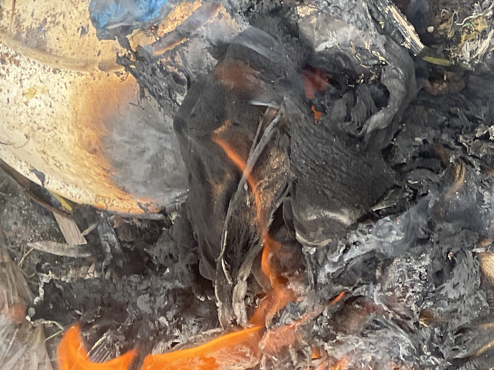 The image depicts a fire burning in Gura Top, Nigeria. The fire consists primarily of trash, including plastic and other unidentifiable debris. The scene is characterized by flames and smoldering remains. The environment is filled with ash and smoke. No people are visible.