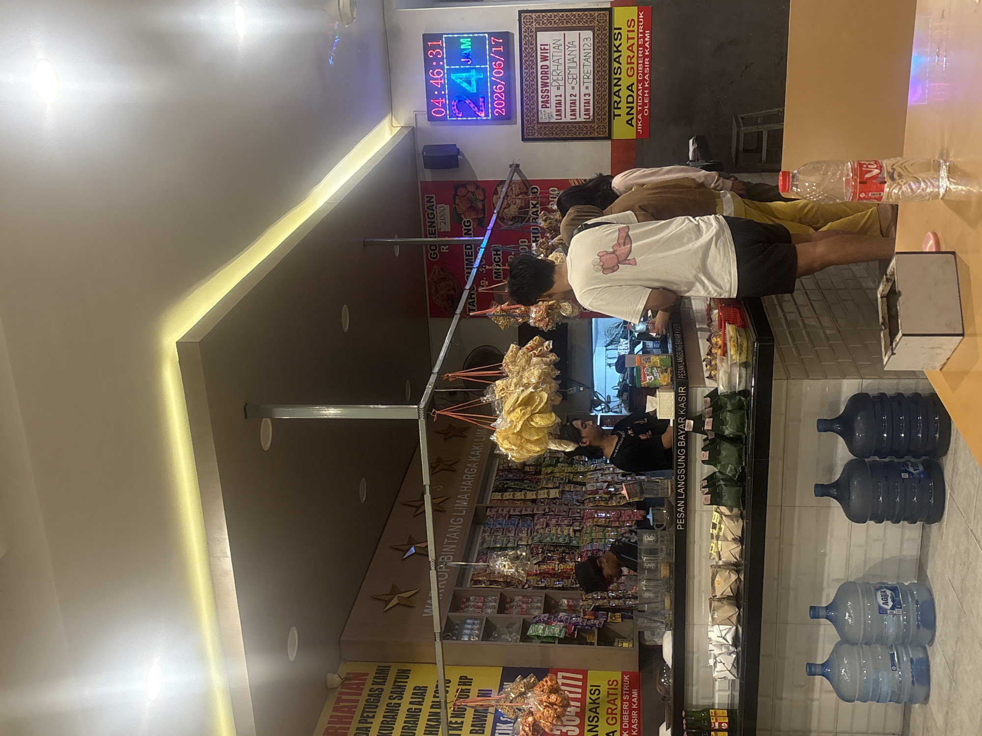 The image depicts the interior of a small shop in Bebekan, Indonesia, likely a convenience store or warung. The scene is brightly lit, with a long counter dominating the foreground and midground, behind which shelves are stocked with various packaged goods, snacks, and drinks. Several customers and staff are visible.
In the midground, two male employees are standing behind the counter. One, wearing a black shirt with "WARKOP" printed on it, appears to be interacting with a customer. The other employee, in a black t-shirt and cap, is also behind the counter. To the right of the counter, three customers are interacting or waiting. The one in the foreground wears a white t-shirt with a pink graphic and black shorts, facing away from the camera. To his right, a person in a brown sweater and yellow pants is facing the counter. Further to the right, a person in a beige shirt is also near the counter.
The counter itself is cluttered with items, including what appear to be beverages in plastic bottles and some sort of food items in red baskets. In front of the counter, on the floor, are several large water jug bottles. A small, square ashtray is on a table in the foreground, next to a bottle of water.
A digital display mounted on the wall to the right shows the time "04:46:31" and the date "2026/06/17". A framed sign below it displays "PASSWORD WIFI" with different access codes for different levels. A yellow sign with red text below that reads "TRANSAKSI ANDA GRATIS" (Your transaction is free).
The walls behind the counter are decorated with various signs, including one with "PERHATIAN" (Attention) and other text in Indonesian, and images of food items. Hanging from the ceiling are displays of bagged snacks and chips. The ceiling features modern lighting, including recessed lights and a strip of yellow LED lighting. The overall impression is a busy, local retail establishment.