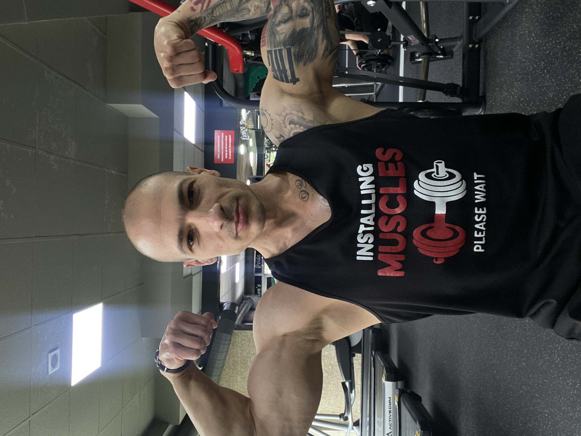A bald man in a black "Installing Muscles" tank top flexes both biceps in what appears to be a gym. He has tattoos on his left arm. The gym has workout equipment visible, and a sign in the background is in Bulgarian, suggesting a location in Pleven, Bulgaria. The environment includes standard gym equipment and overhead lighting.