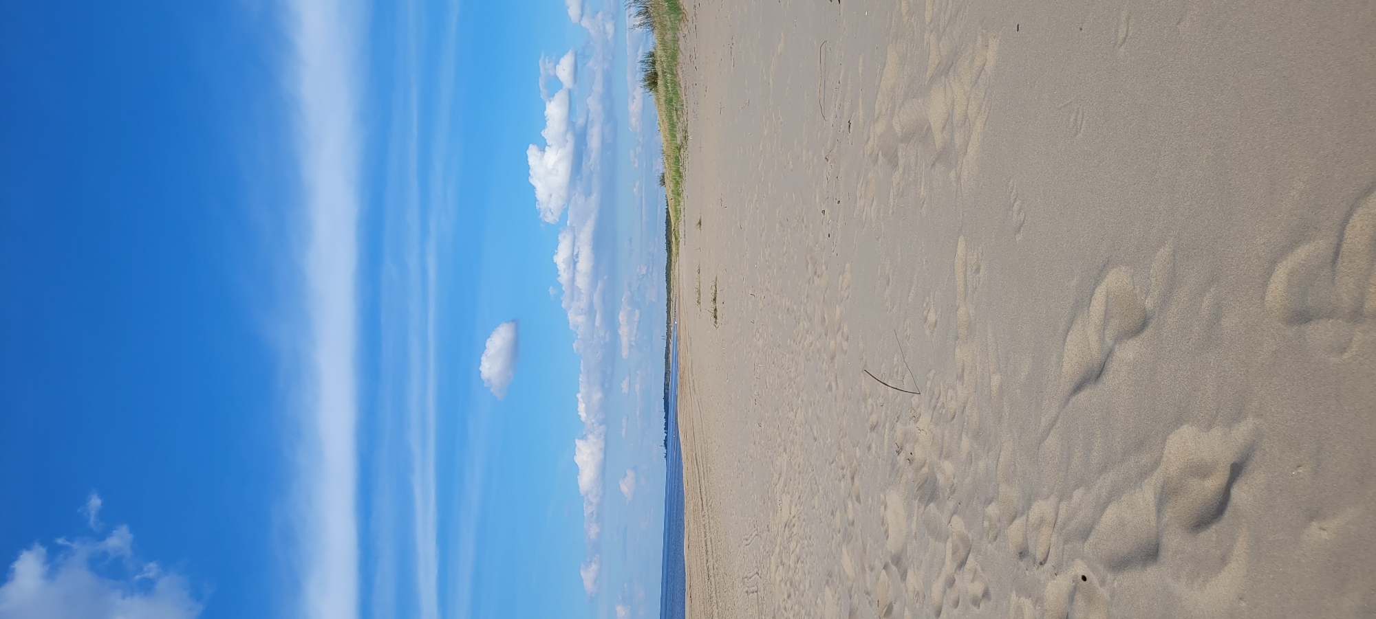 A vertical, eye-level shot of a sandy beach on a clear, blue sky day. The beach stretches from the foreground to a low, grassy dune line in the distance, bordered by the Baltic Sea on the left and a distant, hazy skyline of Riga. The sand is mostly smooth with subtle ripple marks, and a few scattered pieces of dried grass and a single thin stick are visible. The water is calm and blue, meeting the sand in a gentle curve. The sky is a vibrant blue, with streaks of thin, white clouds and a few puffier clouds scattered throughout. The distant Riga skyline, with its industrial structures and faint outlines of buildings, suggests the location is within the city's coastal area.