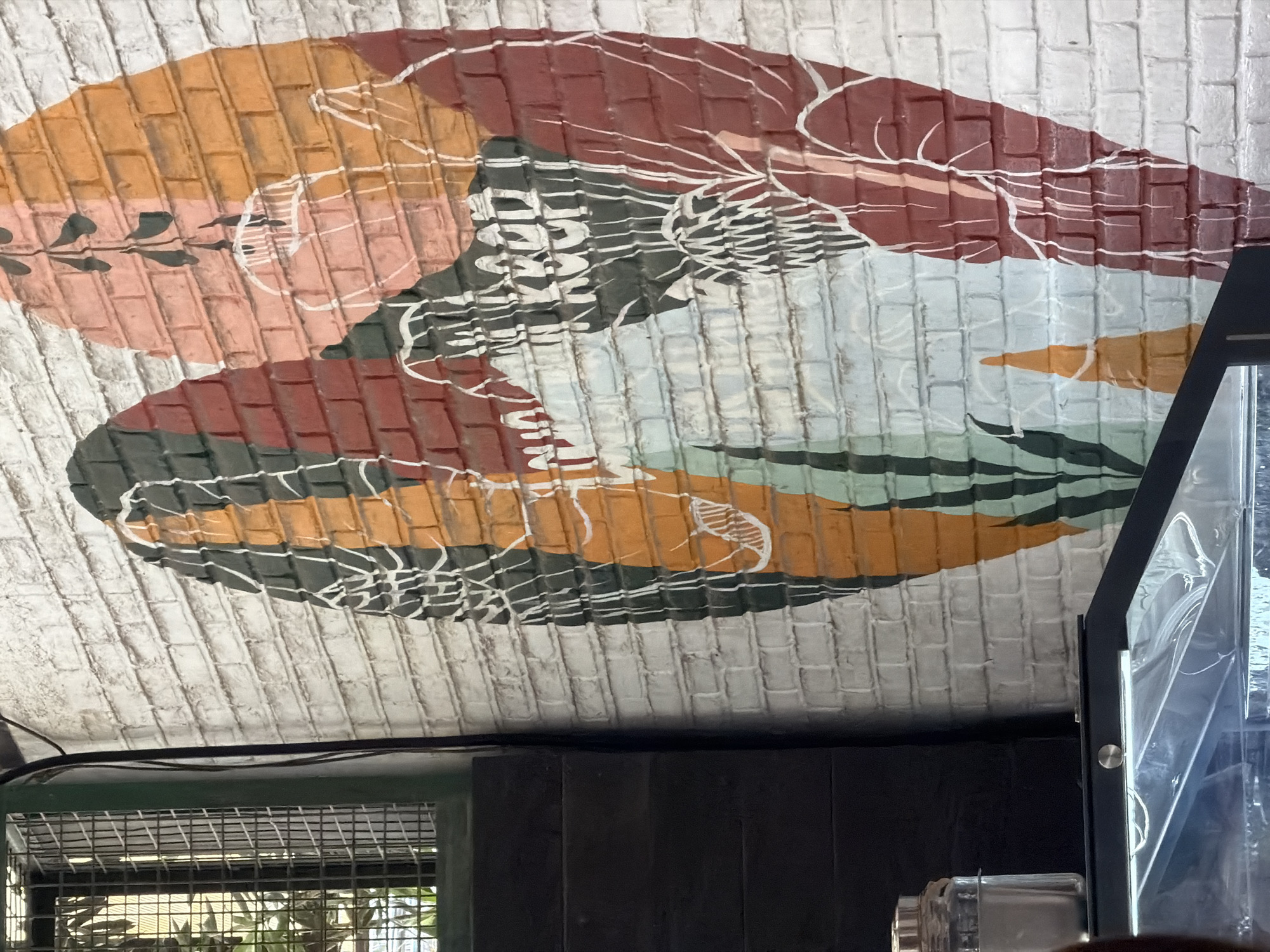 An outdoor mural is painted on a white brick wall in North Kuta, Indonesia. The mural features large, abstract organic shapes in red, orange, green, and black, outlined in white. Some of the shapes appear to be stylized leaves or petals. The mural also contains some white line art resembling veins or branches. To the left of the mural, a dark, industrial-looking structure with a metal grate is visible. Below this structure, a glass display case with a metal frame is partially visible, containing what appears to be a clear plastic lining. A glass jar with a silver lid sits on top of the display case.