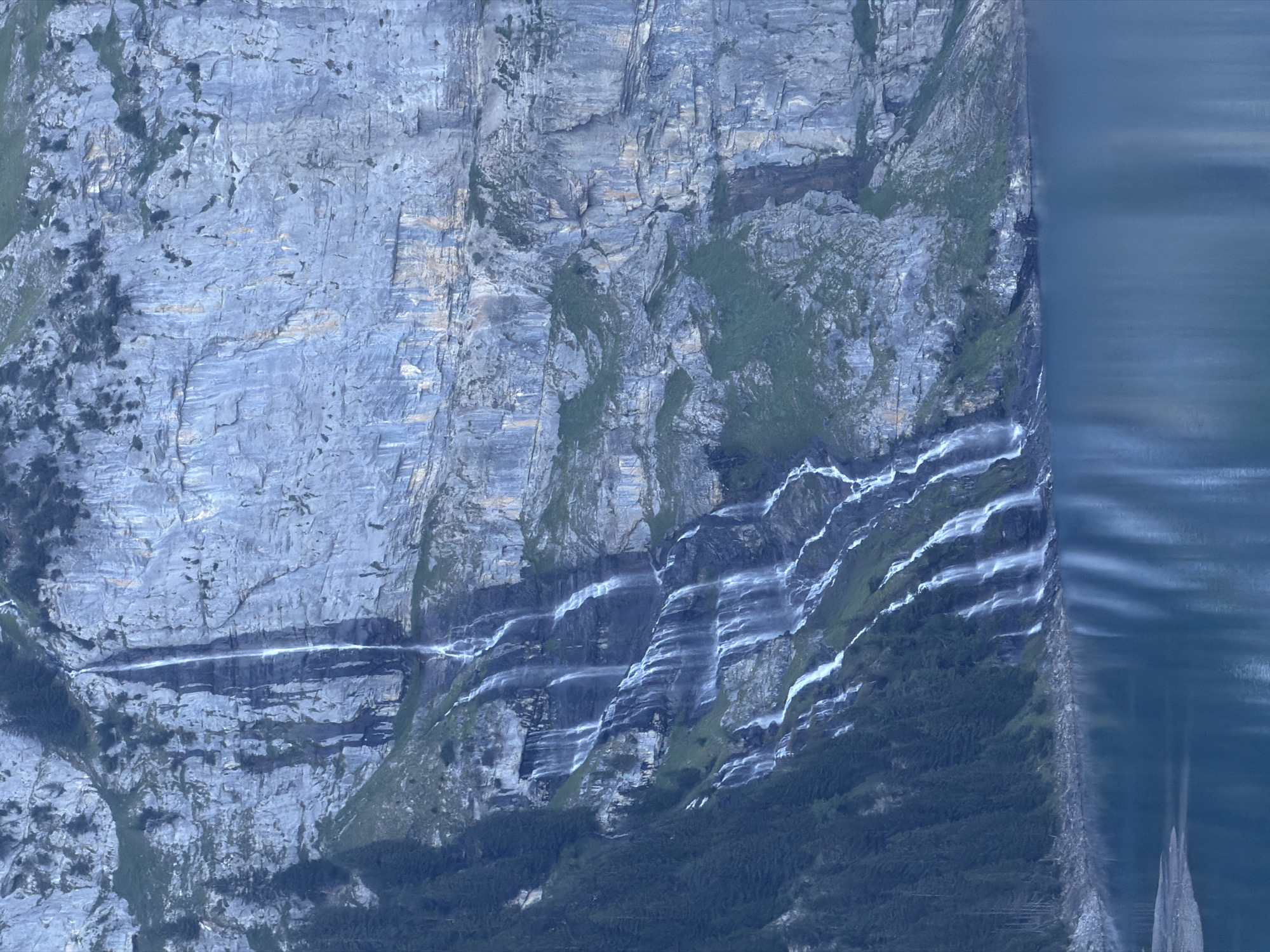 The image shows a large cliff face with multiple waterfalls cascading down its side. The cliff is composed of gray rock with visible strata and some patches of green vegetation. A dense evergreen forest borders the base of the cliff on the left side of the image. The bottom of the image features a body of calm, blue water, likely a lake, reflecting the cliff and waterfalls. The scene is located in Kandersteg, Switzerland, likely at the Oeschinensee lake, renowned for its dramatic alpine scenery and waterfalls.