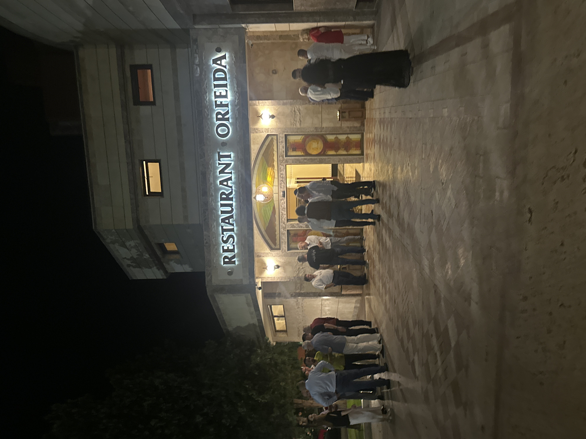 The image depicts a nighttime scene in front of "RESTAURANT ORFEIDA" in Stara Zagora, Bulgaria. The restaurant's name is displayed in large, illuminated letters above the main entrance. The building features a light-colored facade with horizontal textured panels and multiple rectangular windows, some showing interior illumination. The central entrance is adorned with decorative, possibly stained-glass, panels above the double doors and on the doors themselves. Two wall-mounted light fixtures flank the entrance.
Approximately 20-25 individuals are gathered outside the restaurant on a patterned paved surface. The group is predominantly comprised of middle-aged to older men dressed in casual to semi-formal attire, including shirts and trousers. Several smaller clusters of people are engaged in conversation. On the far right, a woman in a long black dress is visible from behind, while another woman in a red top and white trousers stands near the entrance. On the far left, a woman in a dark top and white trousers is also present. A dark-leafed tree is partially visible on the left side of the frame, silhouetted against the dark night sky. The scene is illuminated by the restaurant's signage, internal lights, and exterior sconces, casting shadows on the ground. A small, dark stand or bin is situated near the right side of the entrance.