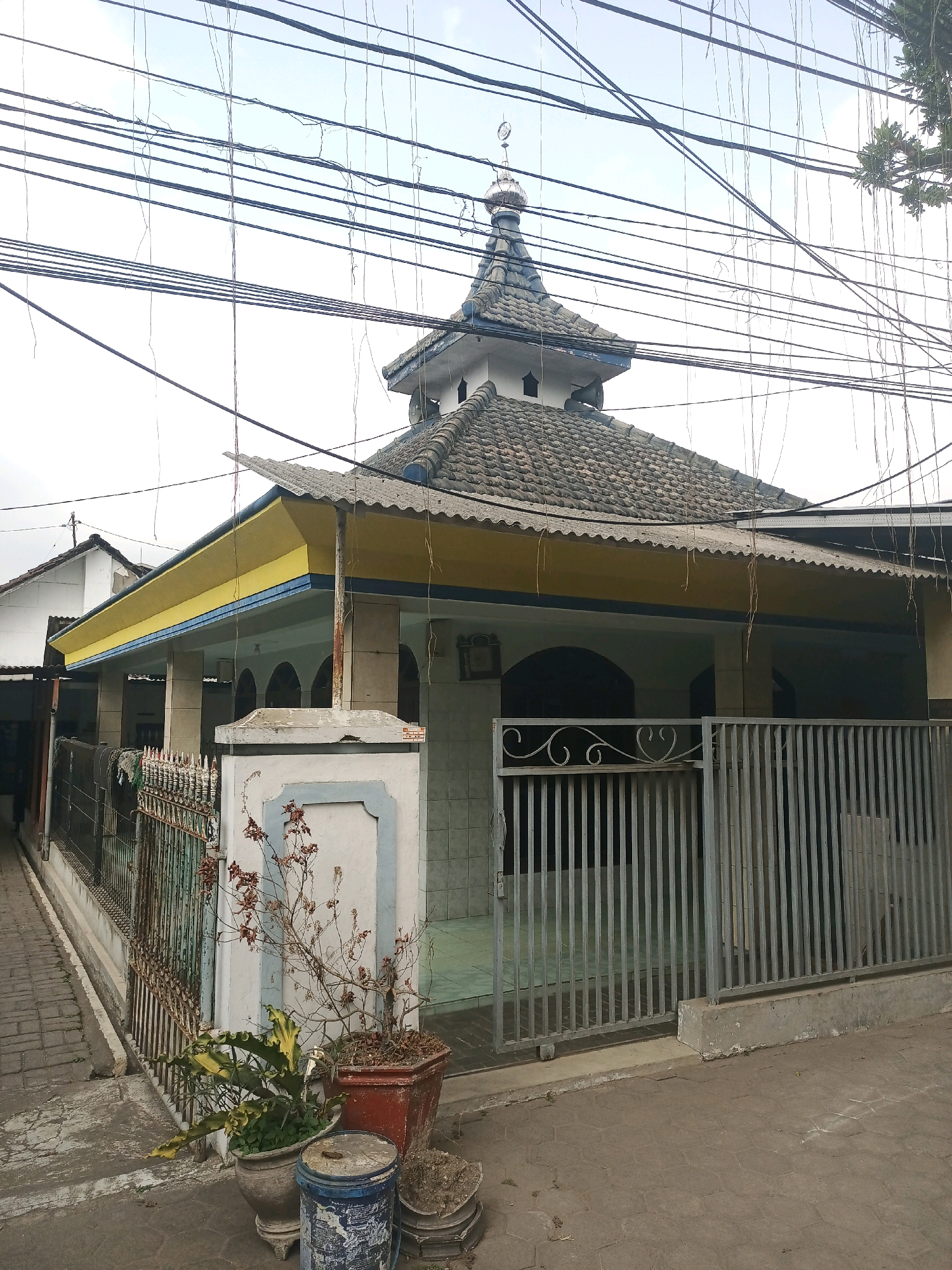 The image depicts a mosque located on a street in Malang, Indonesia. The mosque has a grey tiled roof and a prominent minaret topped with a dome and crescent moon. The building's facade is white with yellow and blue trim under the eaves. A metal fence with a decorative scrollwork gate separates the mosque from the street. Potted plants, including a dried, bare-branched shrub and a leafy green plant, are situated near the entrance. Overhead, a dense network of dark electrical wires crisscrosses the sky, some with hanging vines. The ground is paved with interlocking bricks. To the left of the mosque, a portion of another building with a white roof is visible. The scene is captured under an overcast sky.