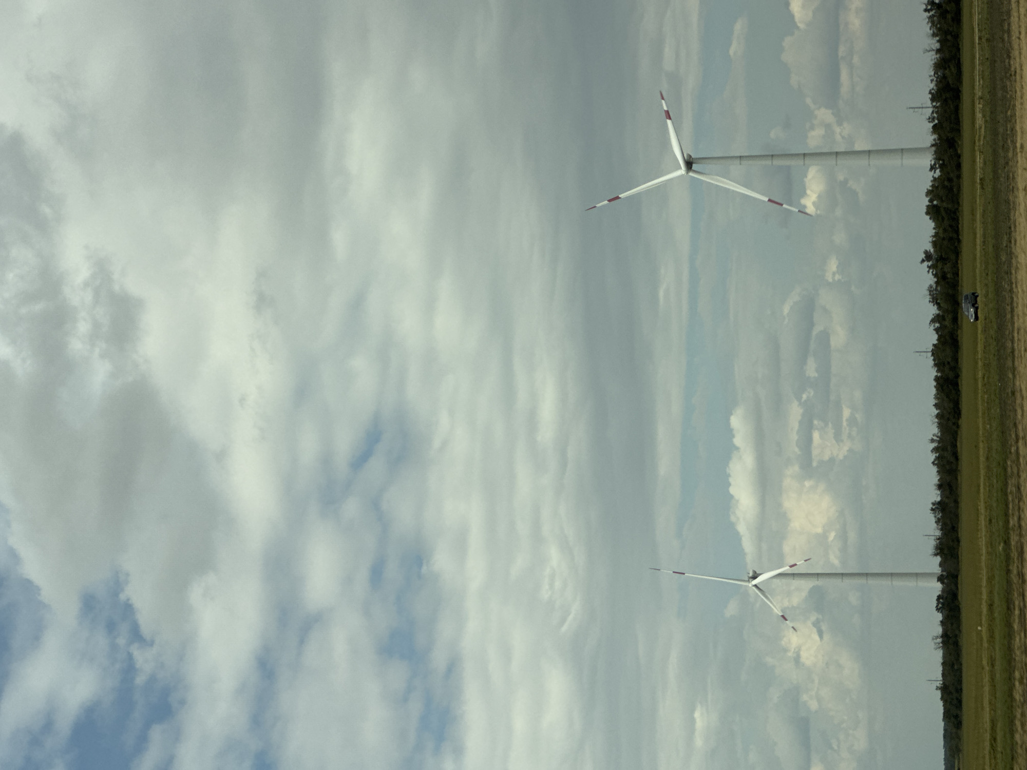 An outdoor, low-angle, full shot captures two white wind turbines in a field under a cloudy sky. The turbine on the left is closer to the viewer, while the turbine on the right is slightly further away. The turbines have three blades, each with red accents on the tips. In the foreground, a green field is visible with a dark-colored SUV parked on the far left. The background features a dense line of green trees, behind which the turbines stand. The sky is filled with scattered clouds of varying gray and white tones. The image appears to be taken from a vehicle traveling through a rural area, possibly on the outskirts of Mayak, Russia.