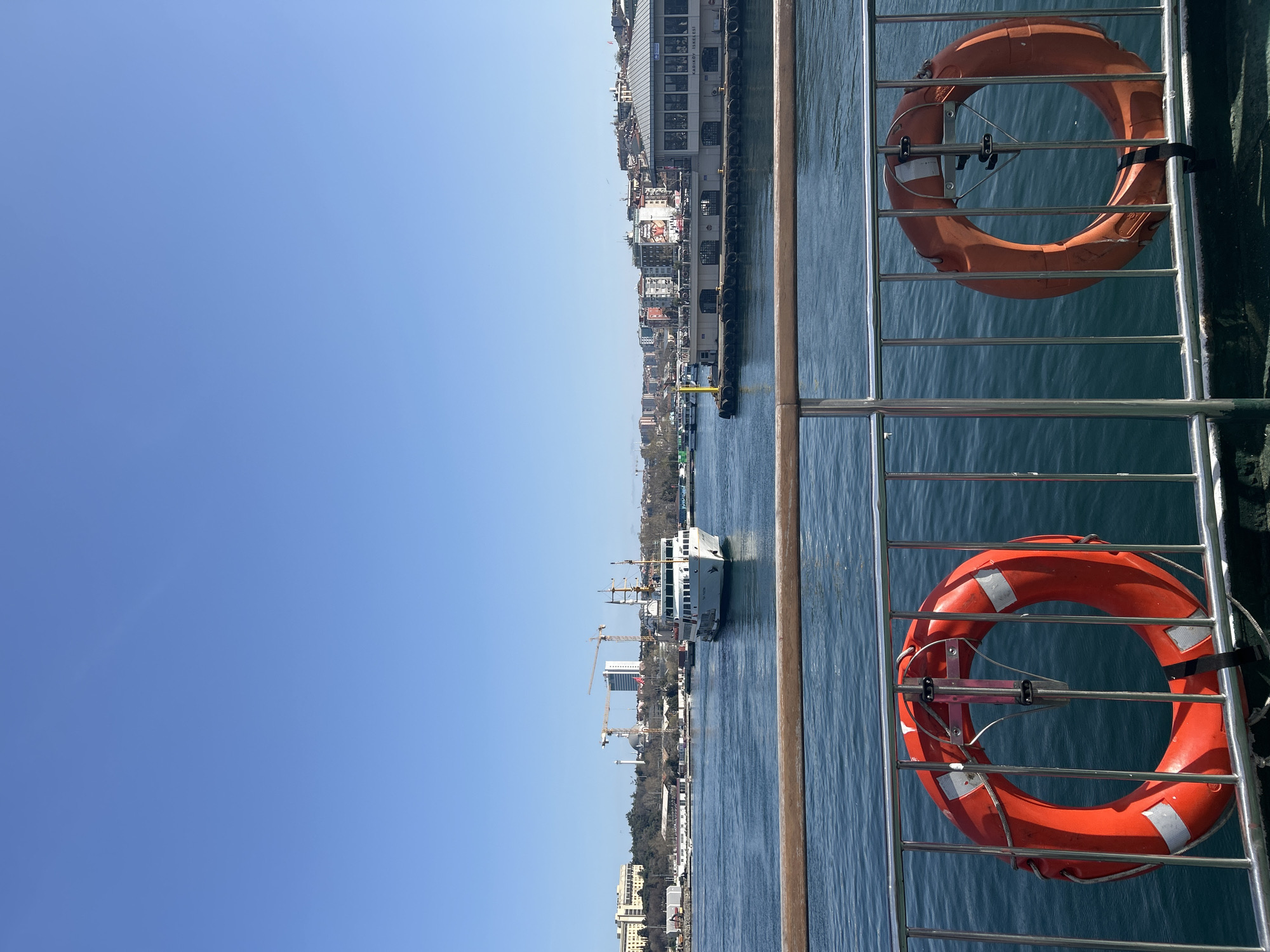 A medium, horizontal shot depicts a view from a boat. In the foreground, two orange life preservers are secured to a metal railing. Beyond the railing, the Bosporus Strait stretches horizontally. A white ferry boat is centered in the water. On the far shore, the Kadıköy cityscape is visible, with buildings of varying heights. Cranes dot the skyline. A clear, blue sky fills the upper portion of the frame.